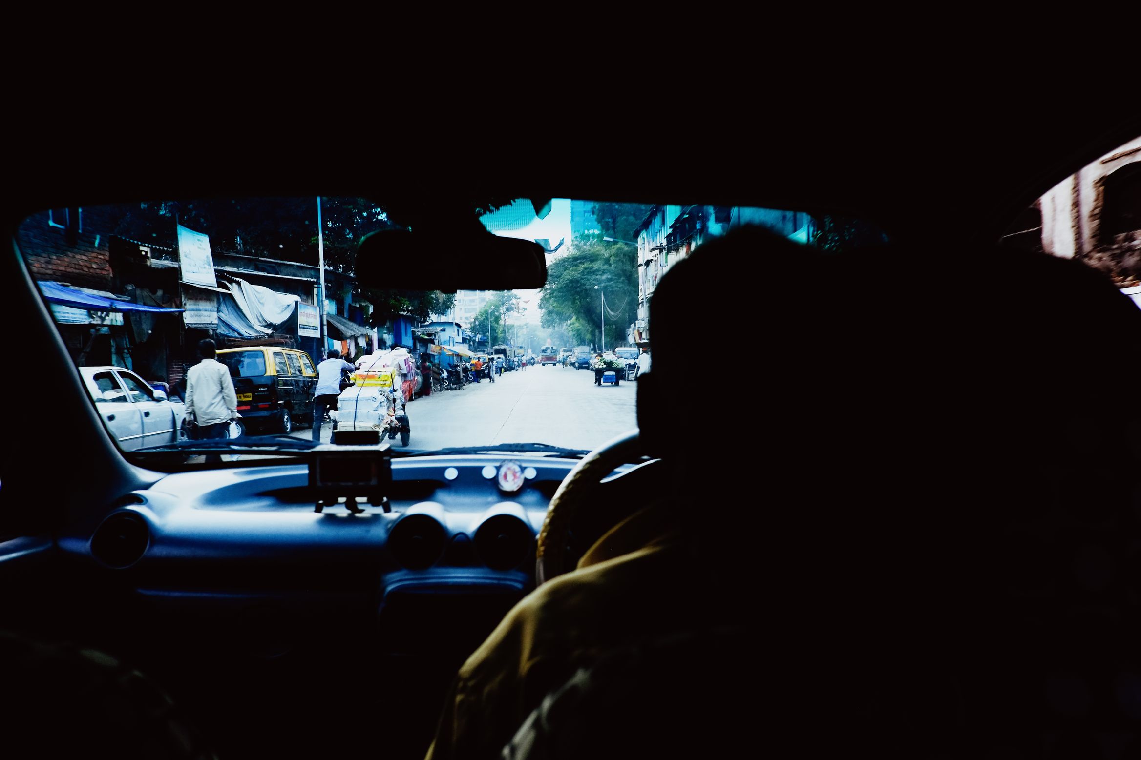 From the back seat of a taxi, the driver in silhouette against a bright street of parked cars and handcarts