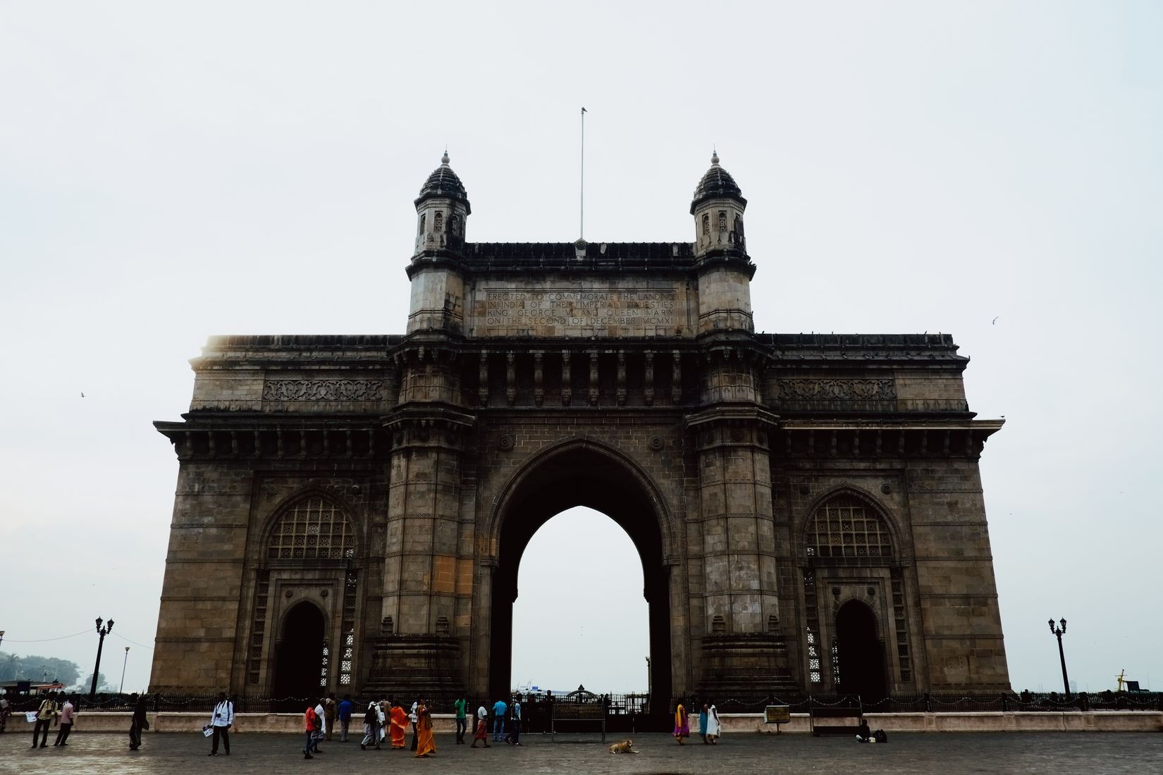 The Gateway of India under flat grey cloud, a scatter of people small along its base