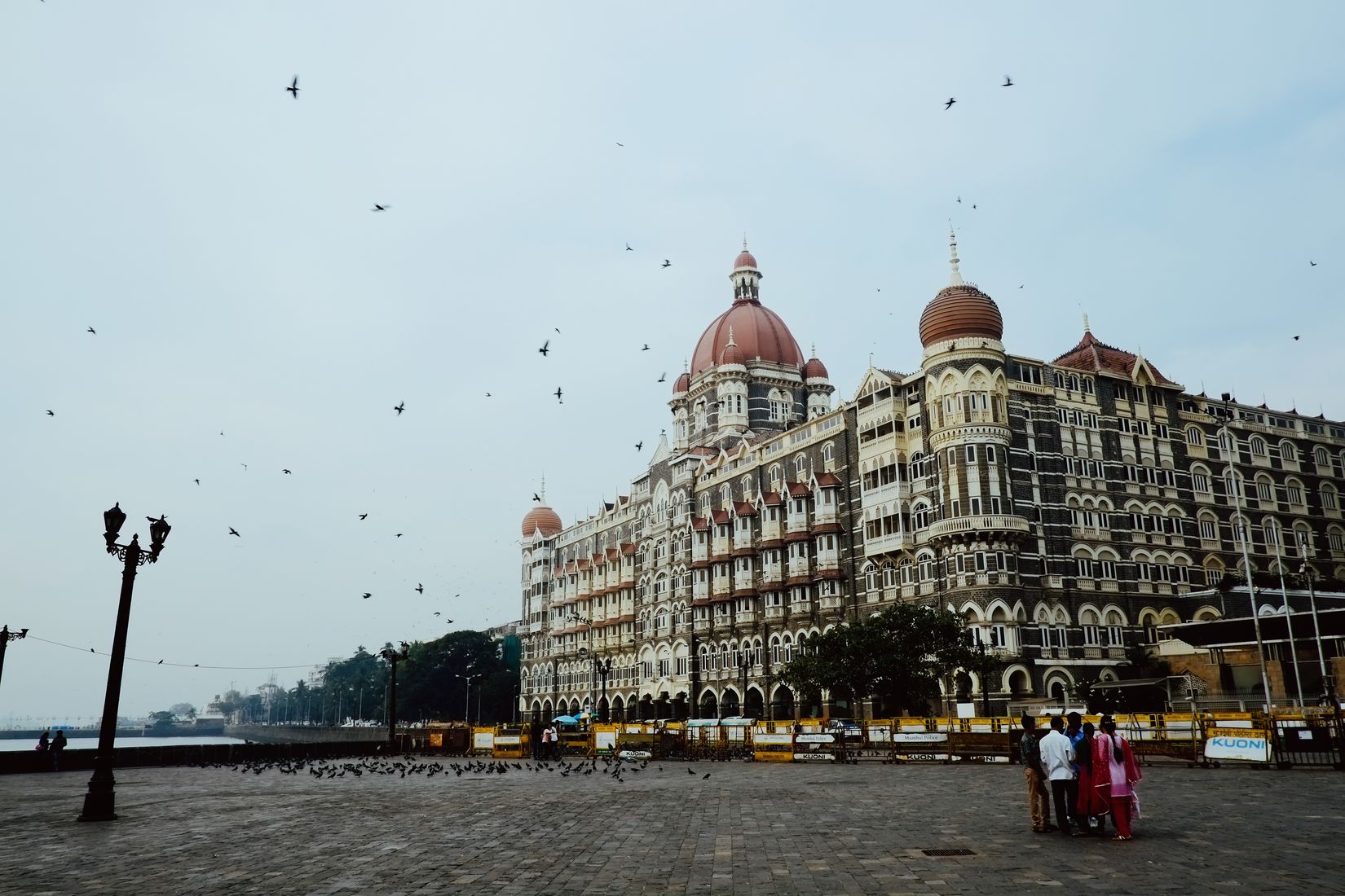 The Taj hotel across an empty plaza with pigeons turning in the air above it, a family of four standing in the foreground