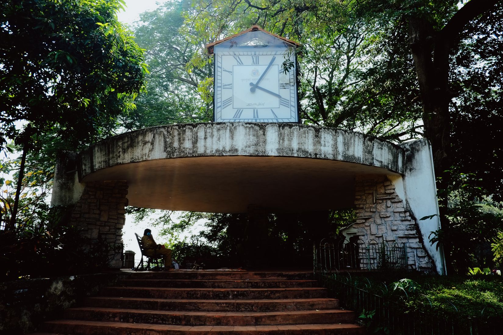 A concrete clock tower on a curved balcony above worn steps, deep in green shade, one person sitting alone underneath