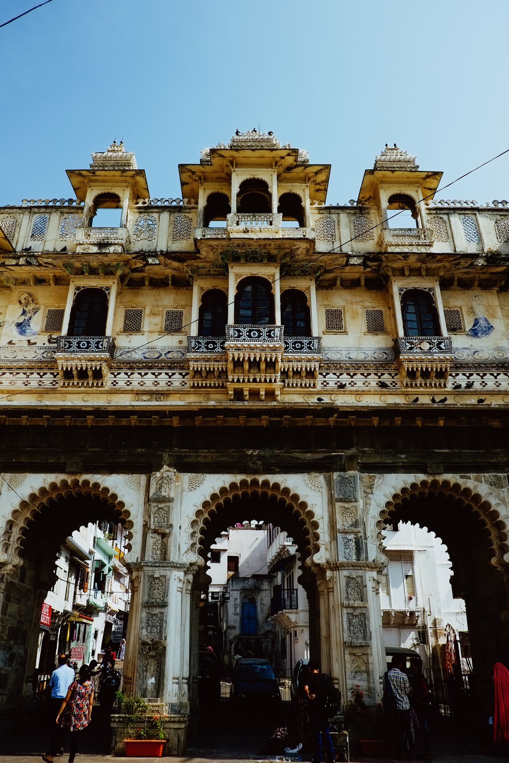 A tiered white gateway with balconies and arched openings, people passing through the arches below into the bazaar