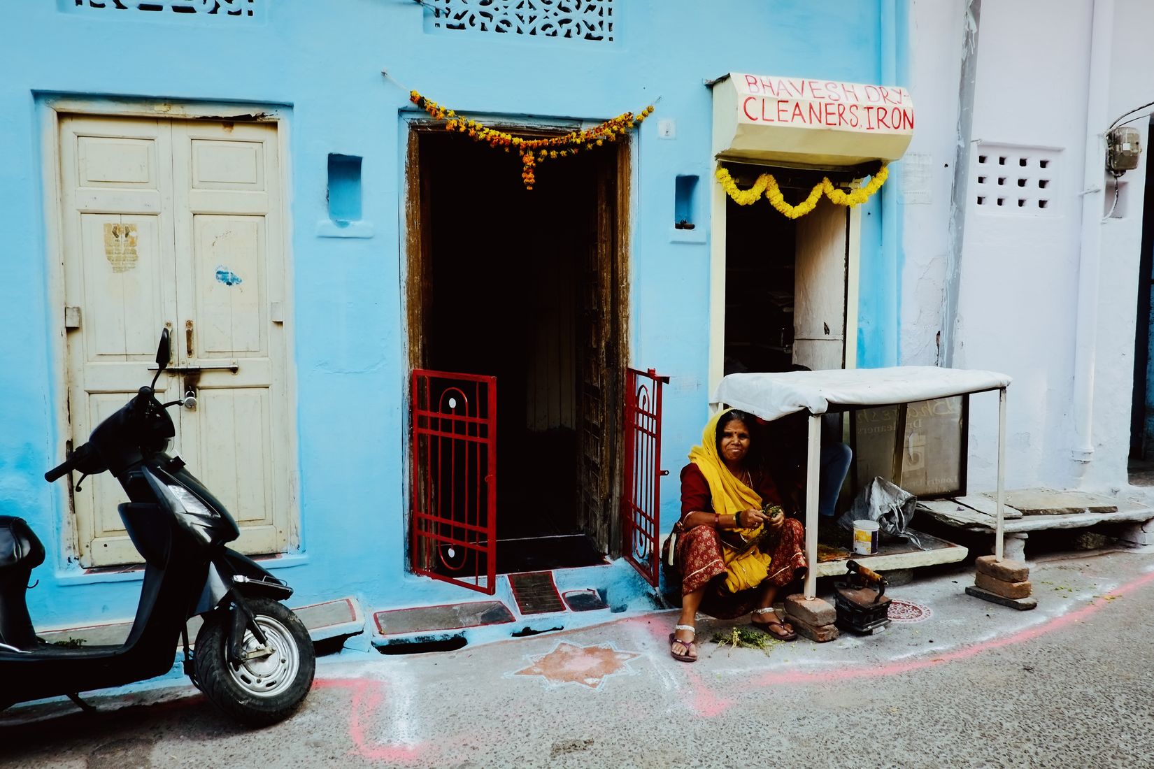 A woman cooking on the pavement outside a bright blue house, scooter parked beside a red gate, marigold garlands over the doors