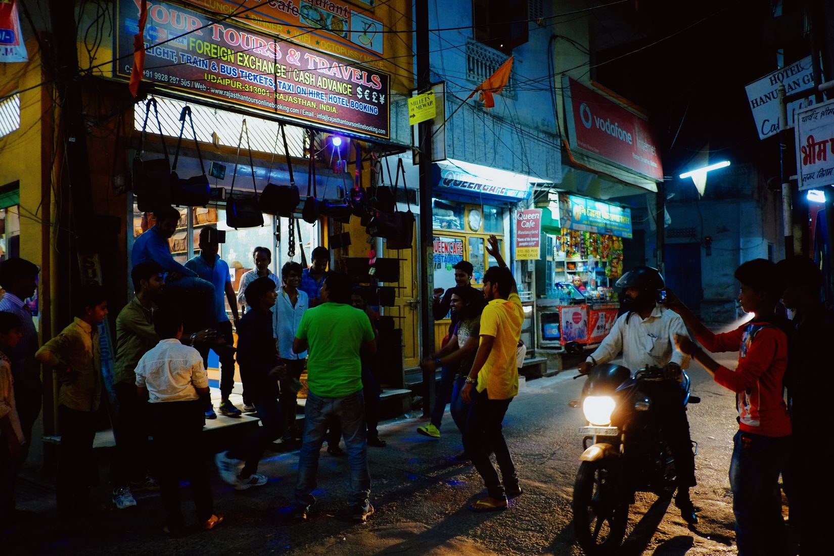 A crowd gathered outside a lit travel agency at night, a motorbike headlight coming through them
