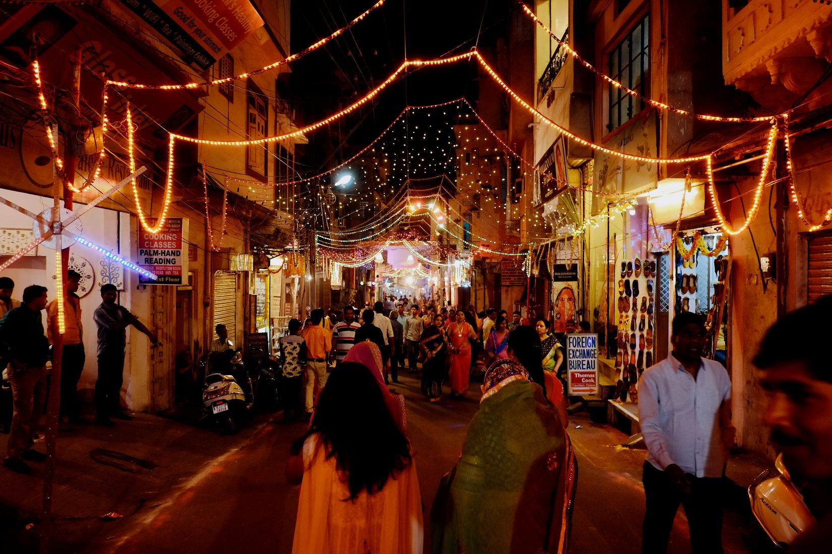 A busy street strung end to end with orange bulbs, women in saris walking towards the camera