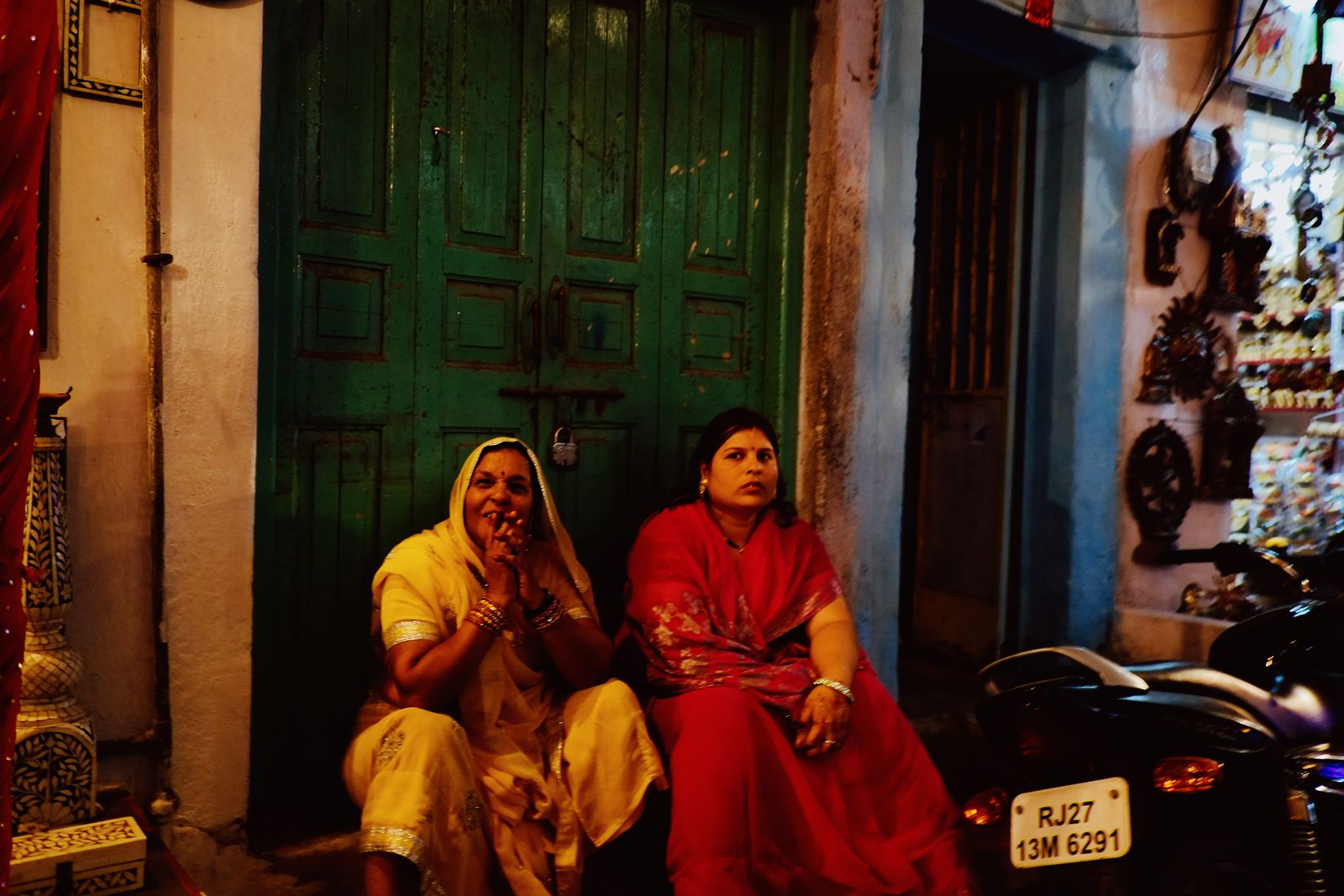 Two women sitting on a step in front of a green door, one in cream and one in red, a rack of souvenirs beside them
