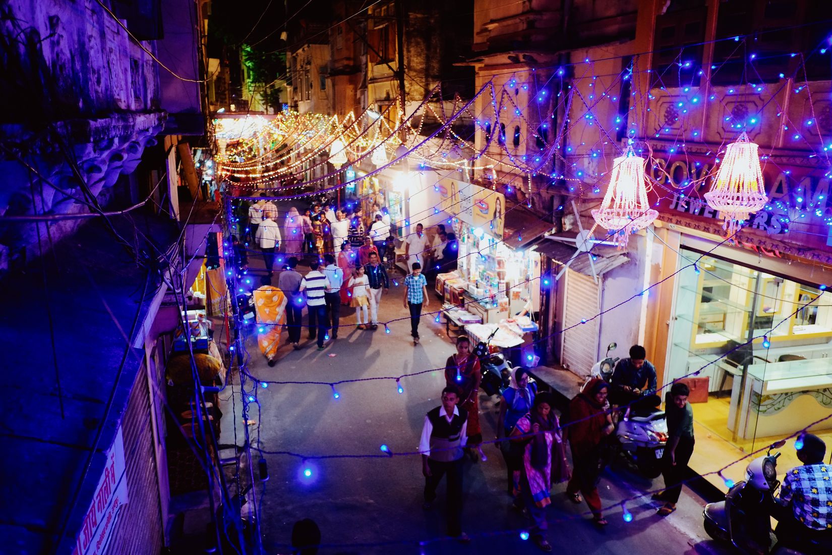 Looking down from a balcony on a street lit blue and white, chandeliers hung outside a jeweller’s