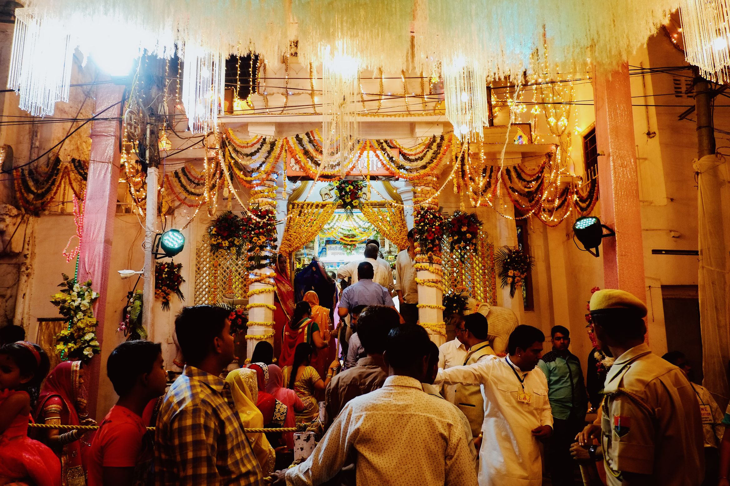 A temple entrance packed with people under heavy marigold garlands and cut-glass lamps, a policeman at the edge of the crowd