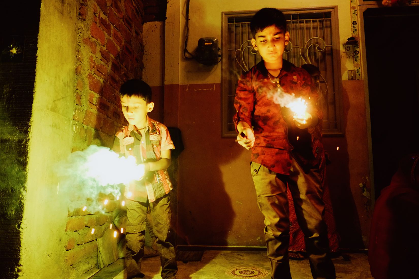 Two boys holding sparklers at arm’s length in a doorway, faces lit hard from below