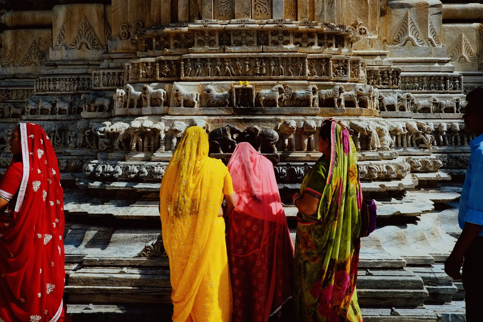 Four women in red, yellow, pink and green saris standing at the carved base of a temple