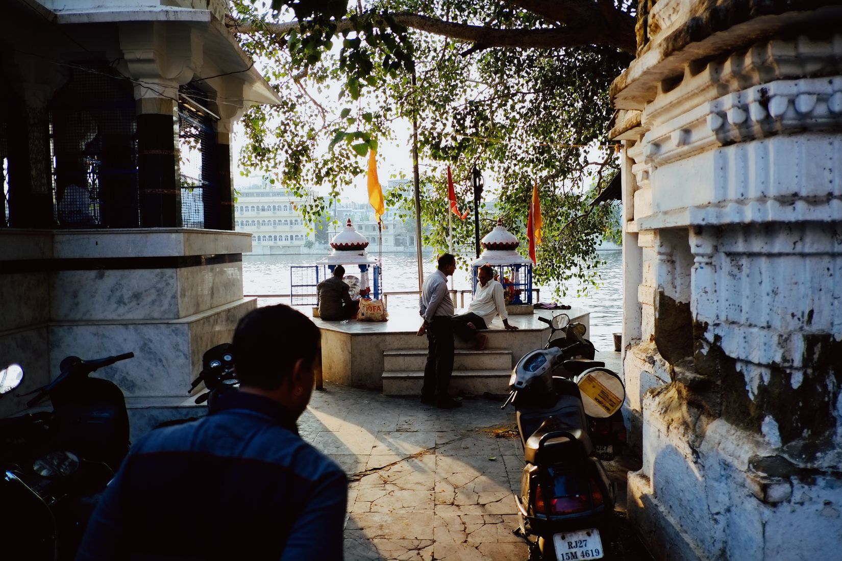 Looking out from a shaded temple porch to the water, orange flags on the ghat and scooters parked across the flagstones