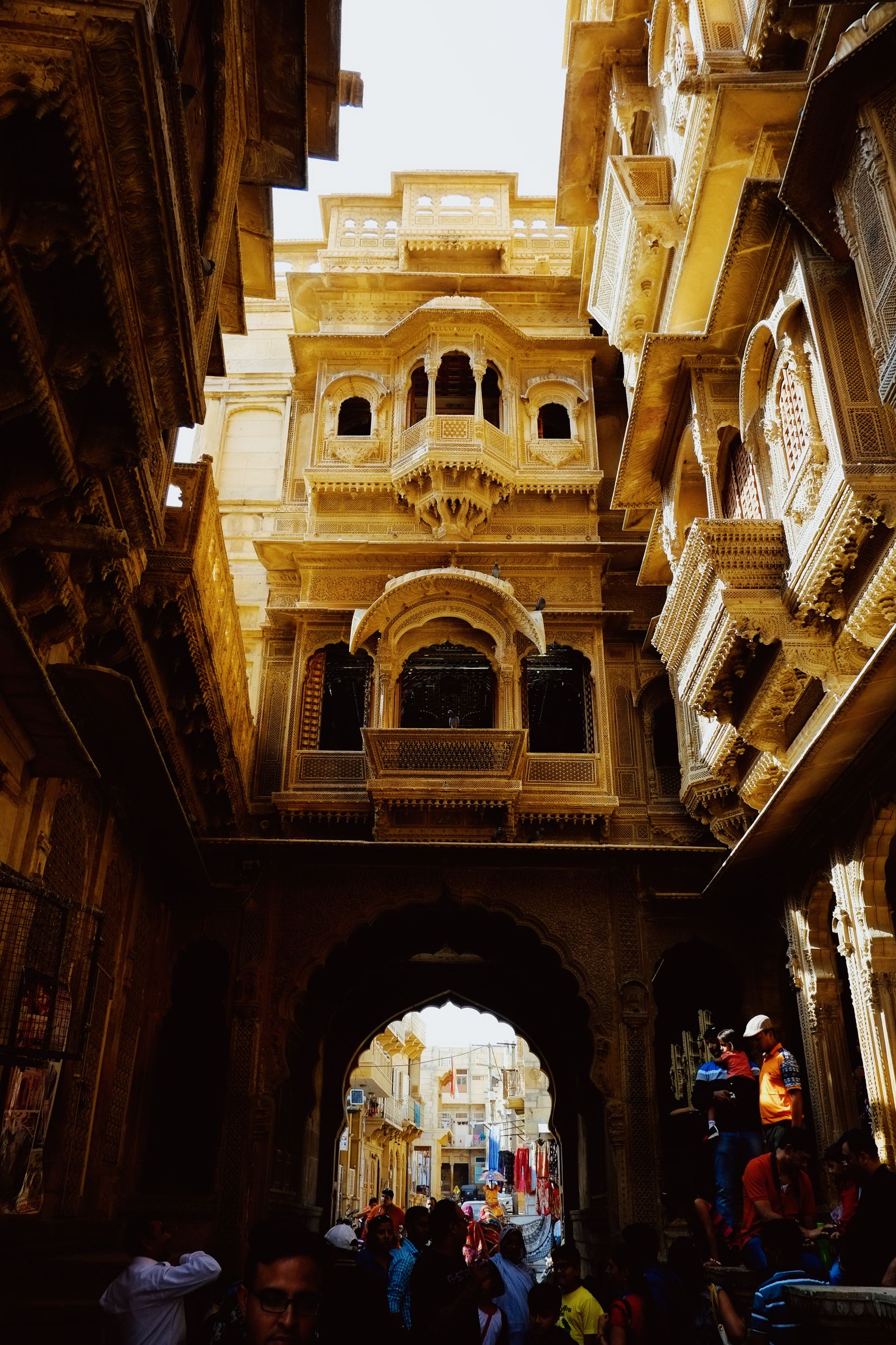 A haveli façade of tiered projecting windows above an archway, people passing underneath into the light