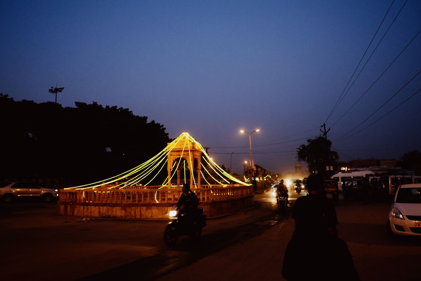 A roundabout monument outlined in yellow bulbs at dusk, motorbikes and cars going round it