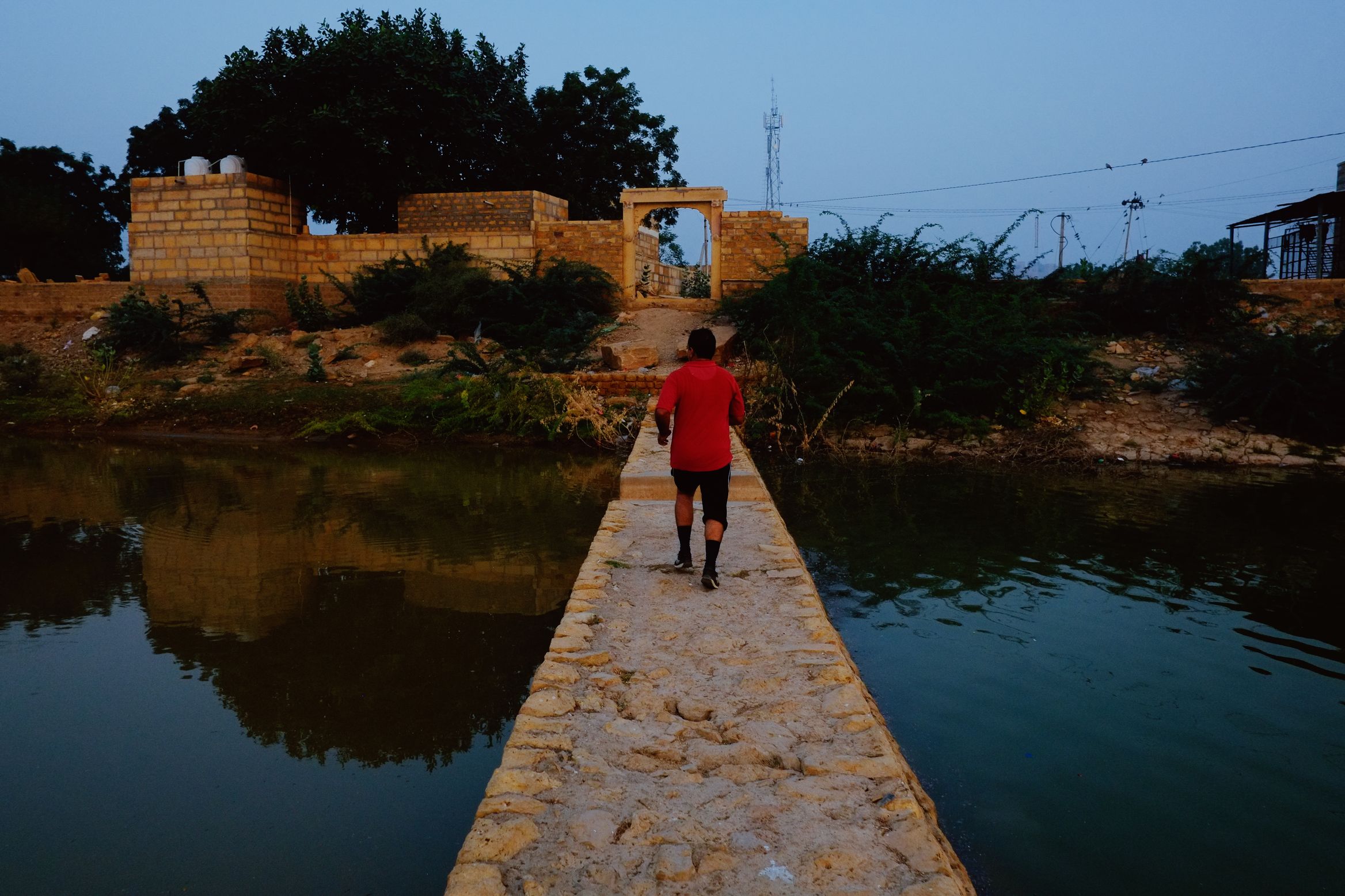 A figure in a red shirt walking a narrow stone causeway across water towards a low sandstone gateway