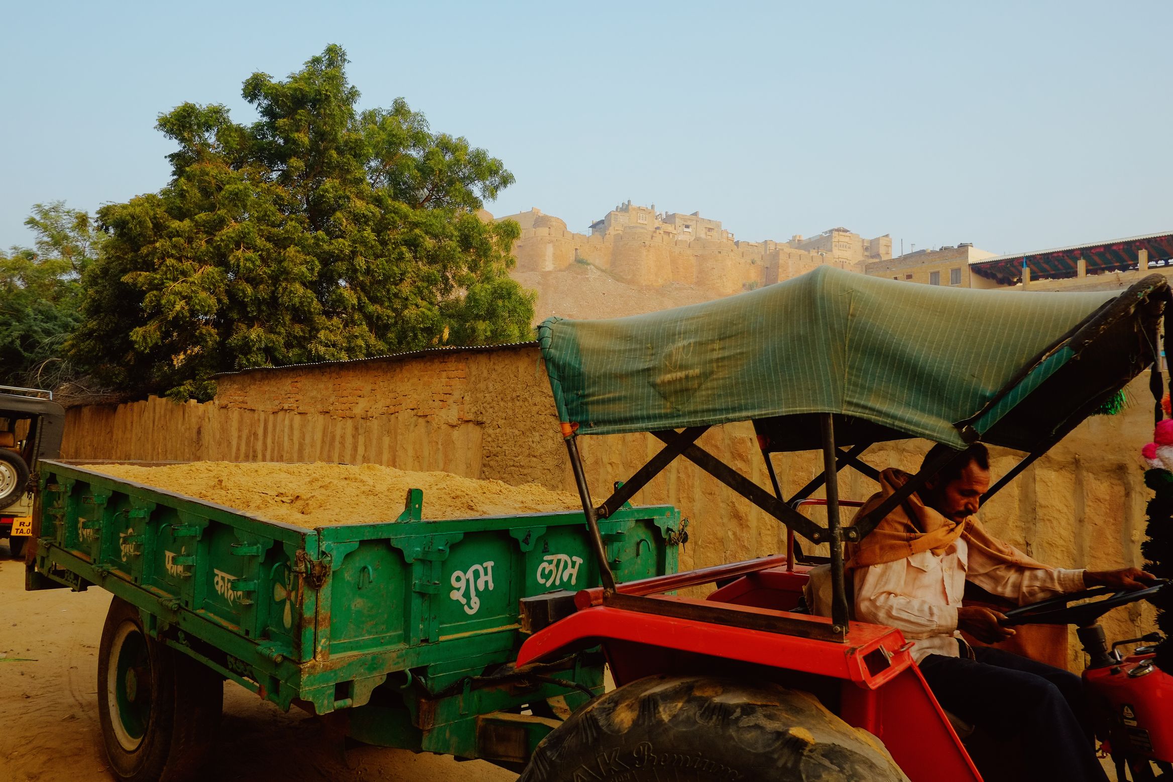 A man driving a red tractor and green trailer loaded with sand, the fort walls on the ridge behind him