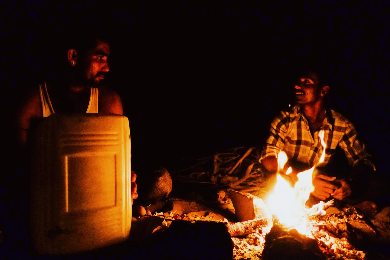 Two of the camel men talking across a low fire, faces lit orange, a water drum between them