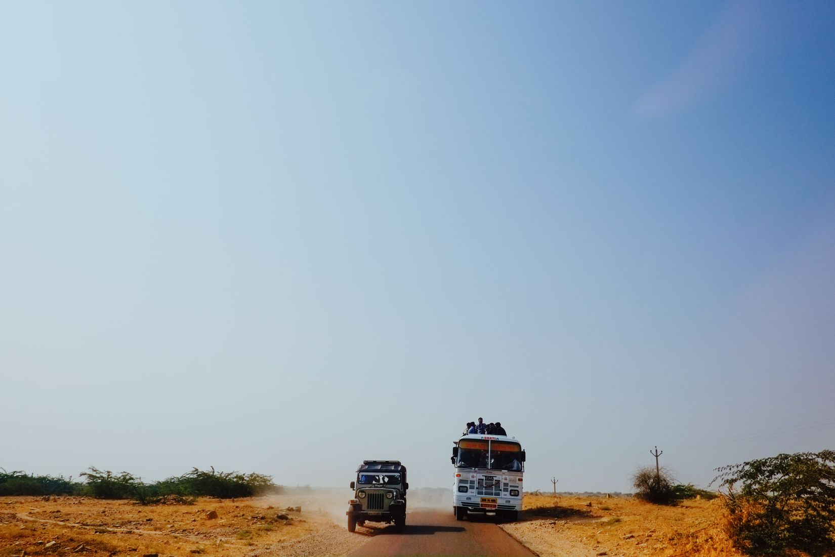 A jeep and a painted bus coming the other way down an empty desert road, dust behind them and men riding on the bus roof