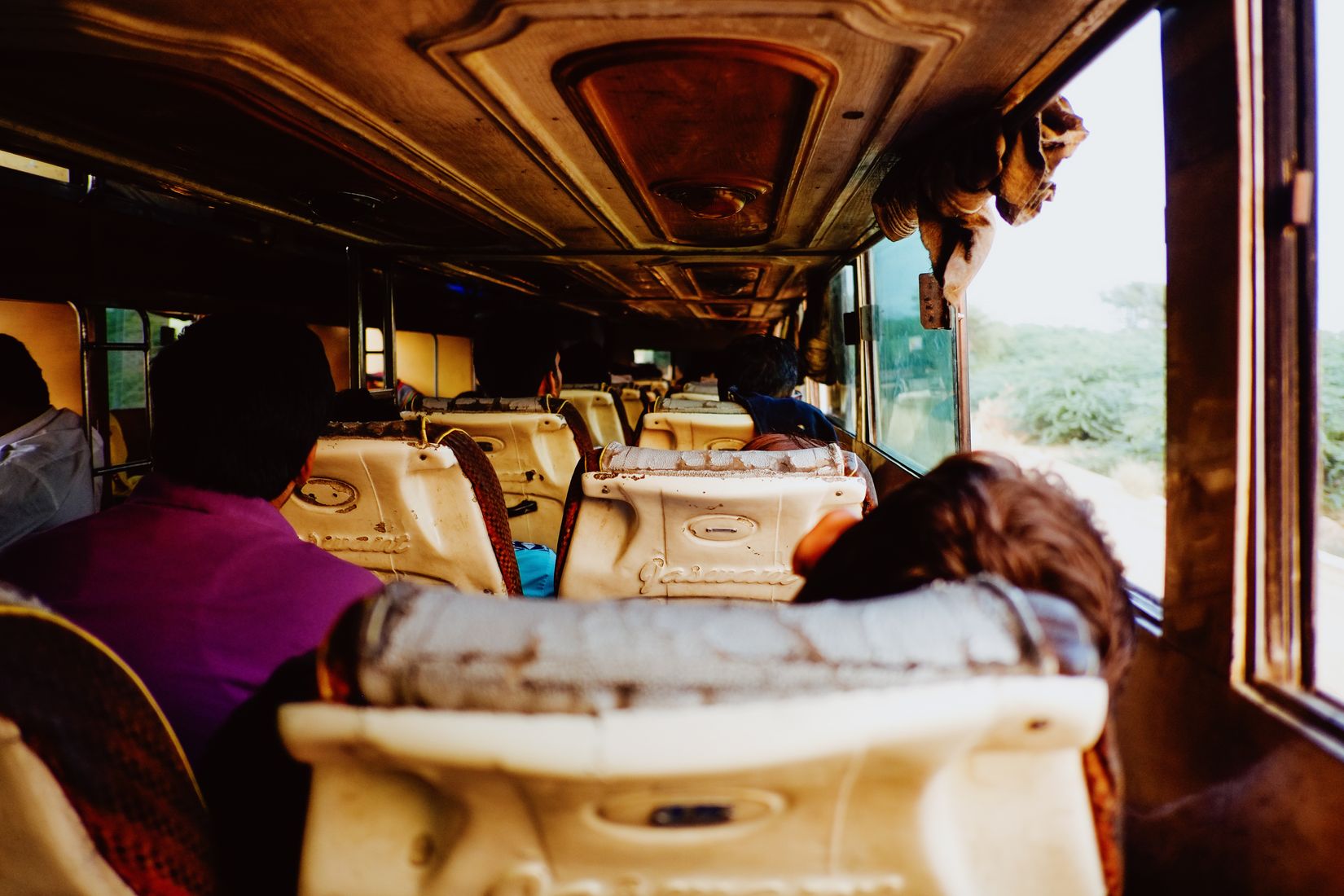Inside a near-empty bus, cream vinyl seats and a bedroll on the rack, dry country going past the window