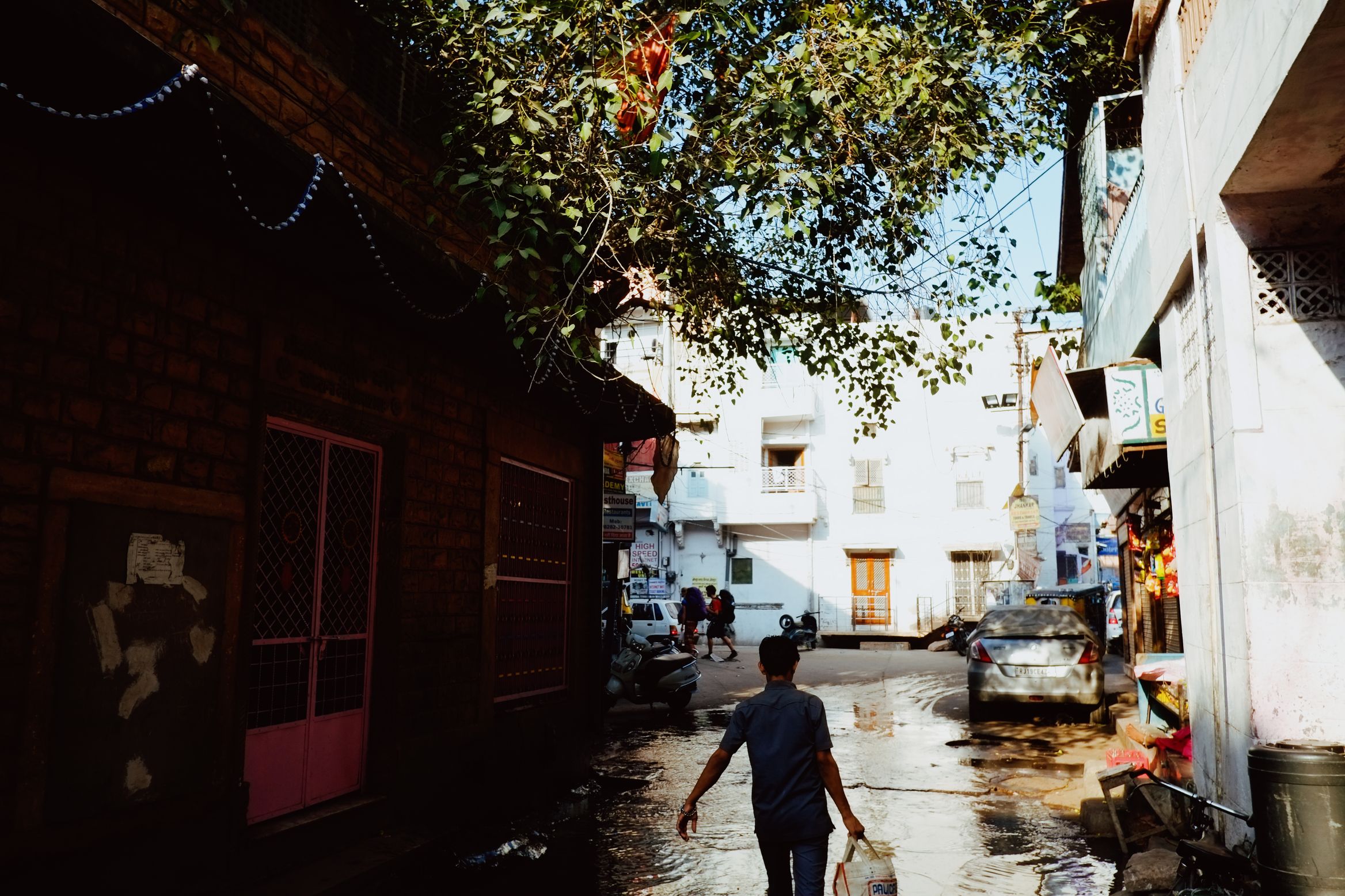 A boy walking away down a wet lane between a brick house and blue-washed walls, carrying a bag