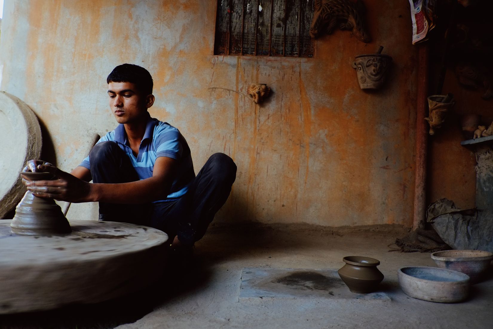 A young potter sitting cross-legged at a stone wheel in an ochre room, shaping a pot between his hands