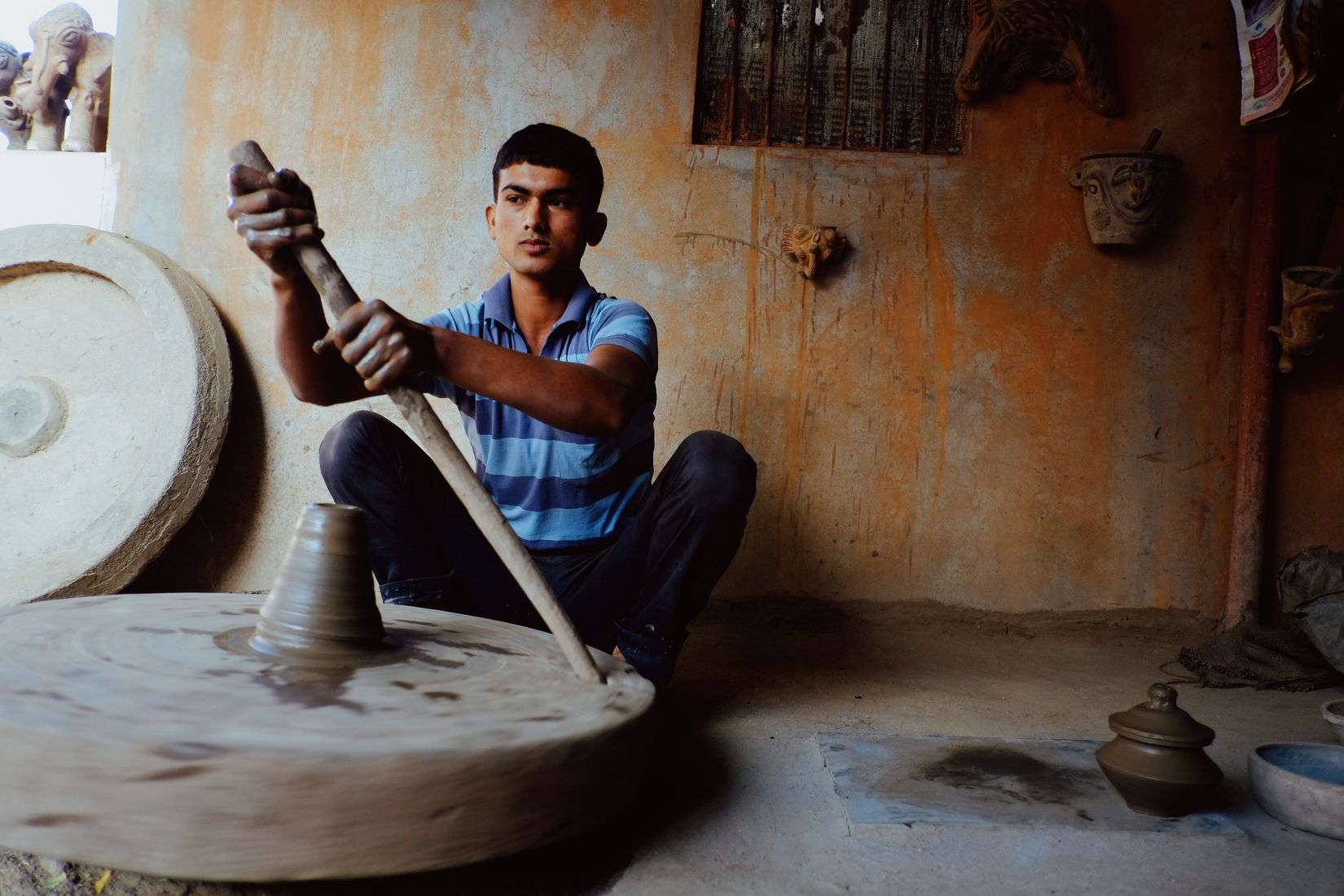 The same potter driving the heavy wheel round with a wooden stick, looking straight at the camera
