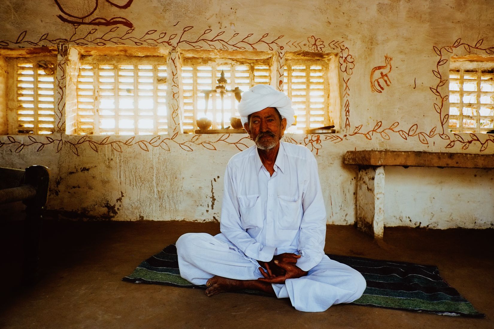An older man in white sitting cross-legged on a rug in front of a wall painted with red vine patterns, hands folded