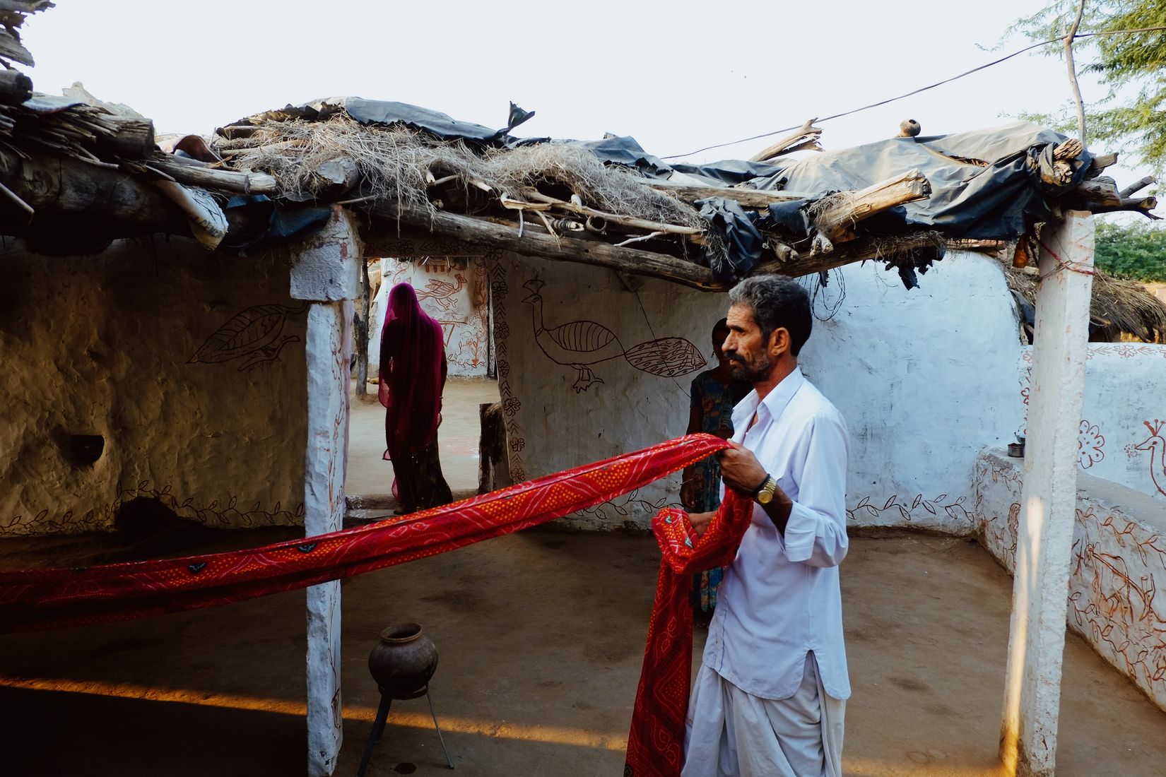 A man holding out a length of red cloth outside a mud-walled house, a woman in red standing in the doorway behind