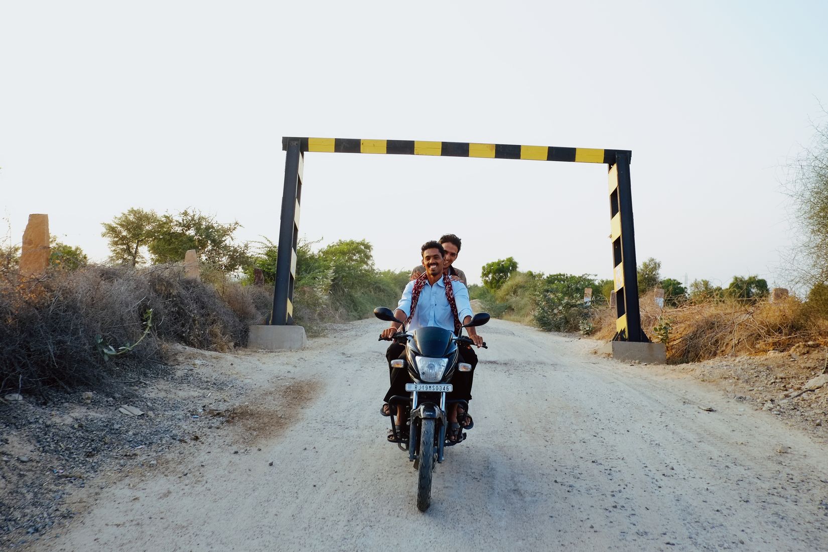 Two men riding one motorbike towards the camera down a white gravel track, under a yellow and black height barrier
