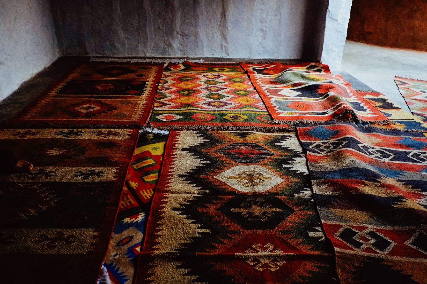Woven rugs laid overlapping across a stone floor in raking light, reds and blues in diamond patterns