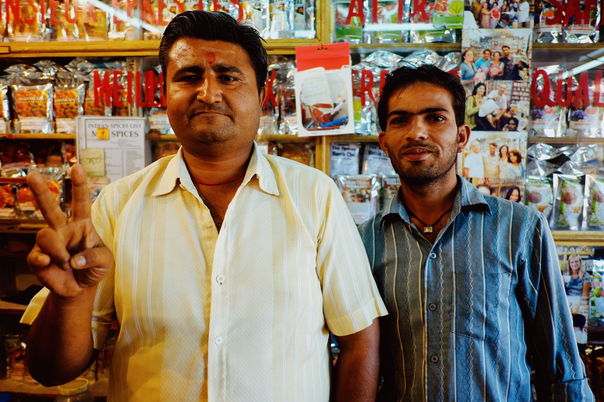 Two men behind the counter of a spice shop, the older one giving a peace sign, packets and film posters stacked behind them