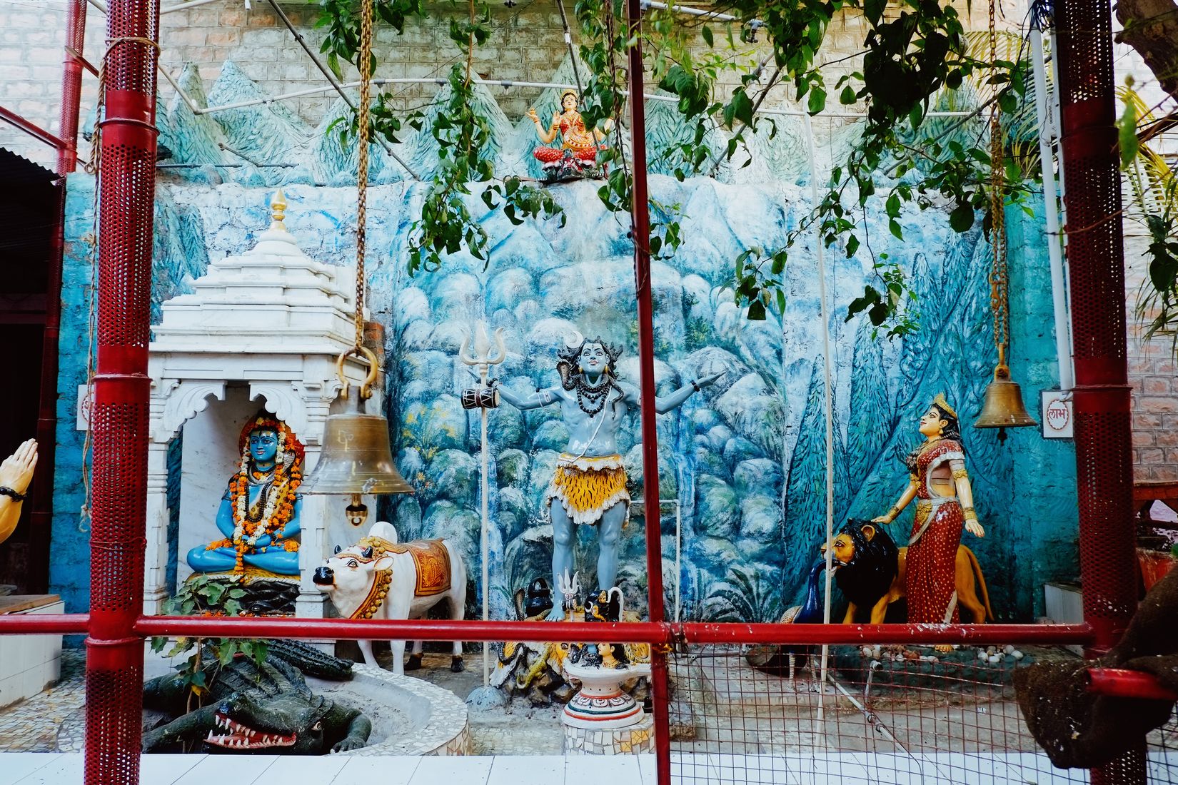 A painted shrine behind red railings — Shiva on a blue mountain with a bull, a bell hanging in front