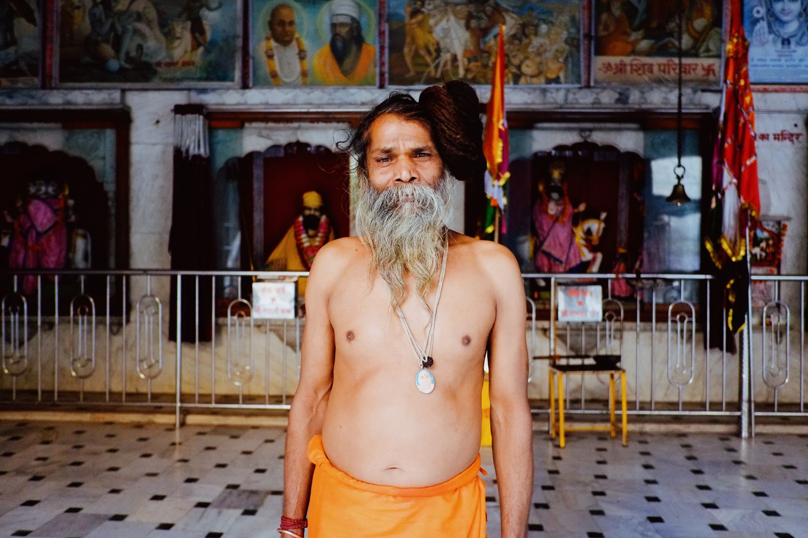 A sadhu in an orange dhoti standing bare-chested in a temple courtyard, grey beard to his chest, looking level at the camera
