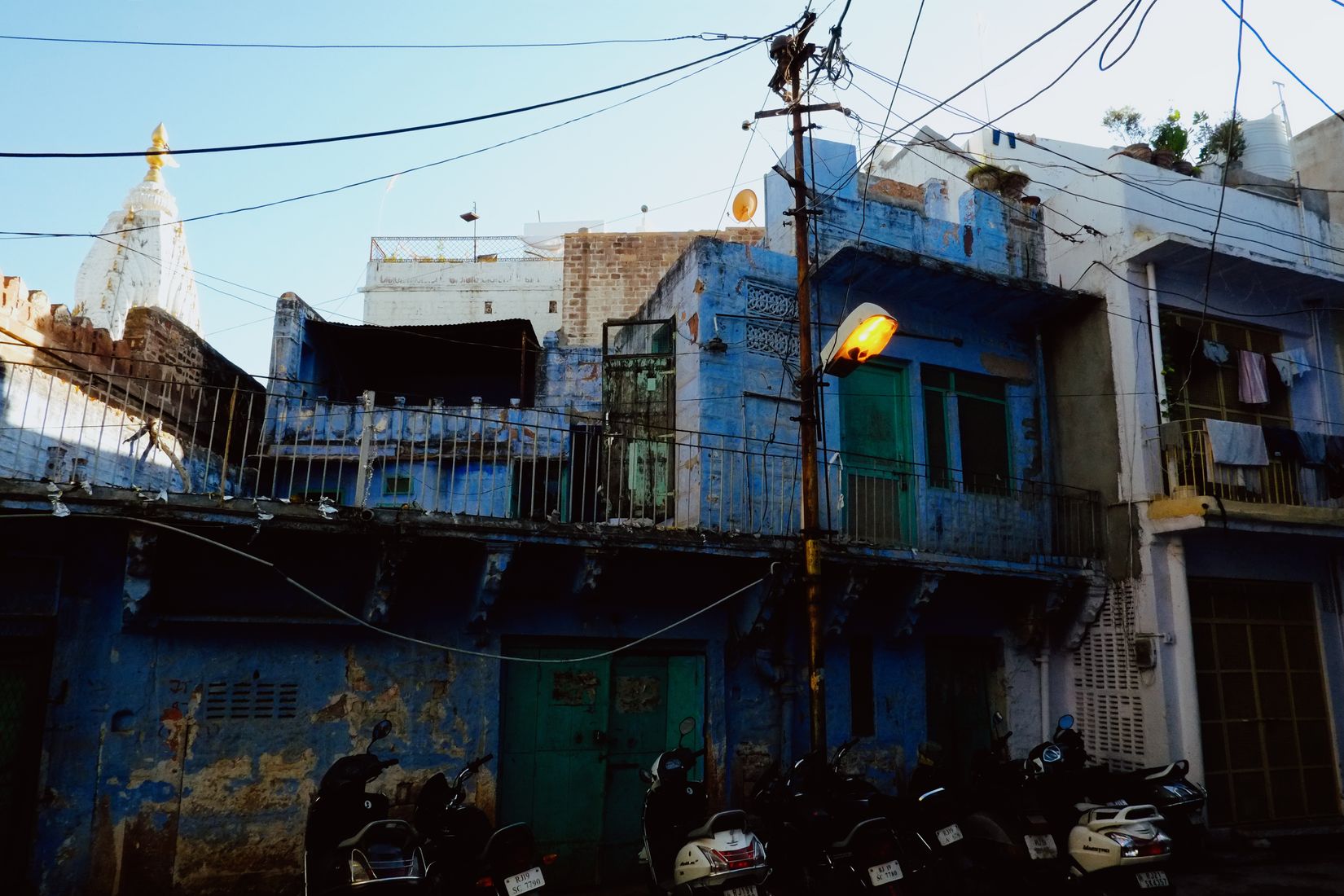 Blue-washed houses with peeling paint and green shutters, a white temple spire rising behind, scooters lined up along the wall