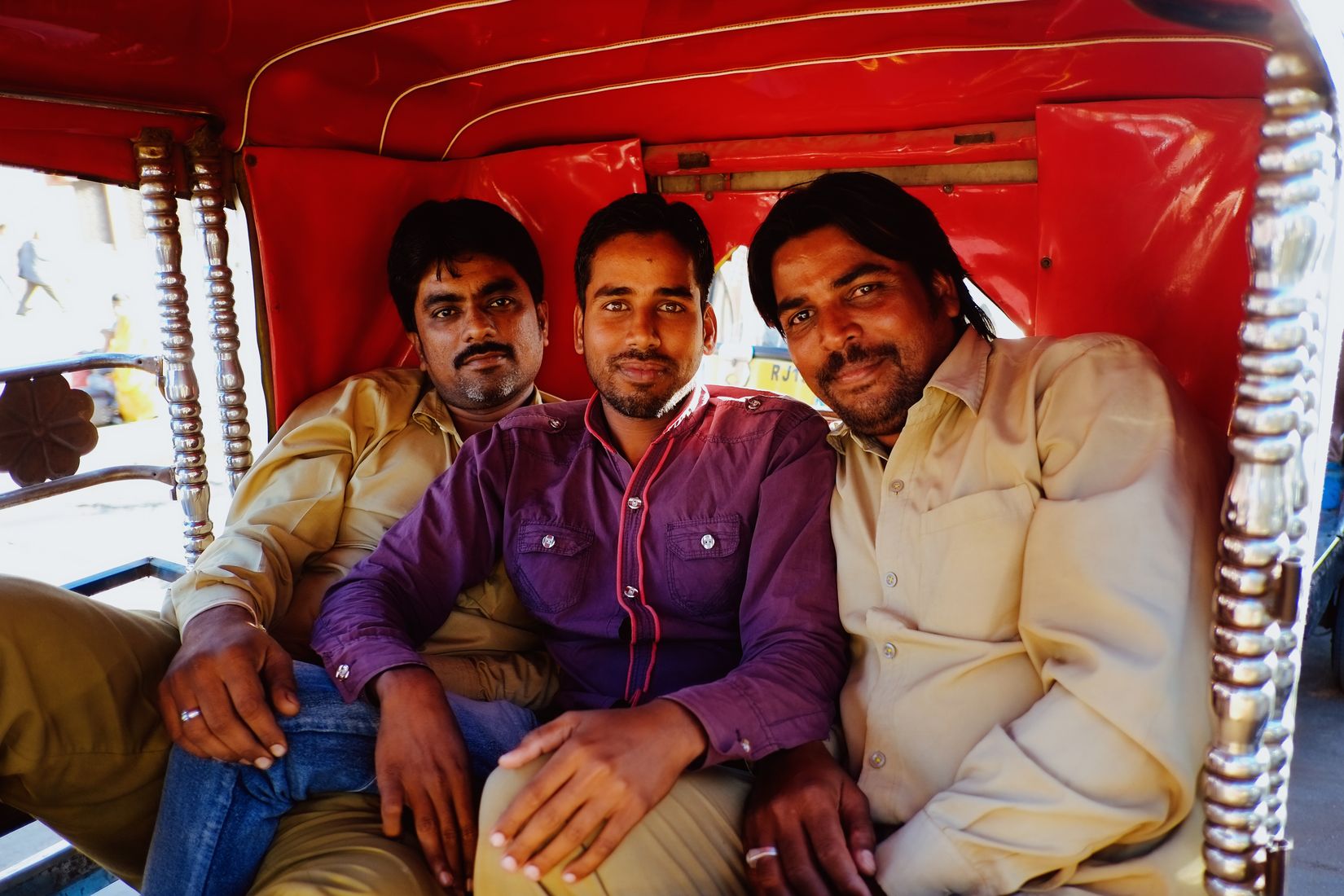 Three men squeezed onto the red bench seat of an auto-rickshaw, all looking into the lens