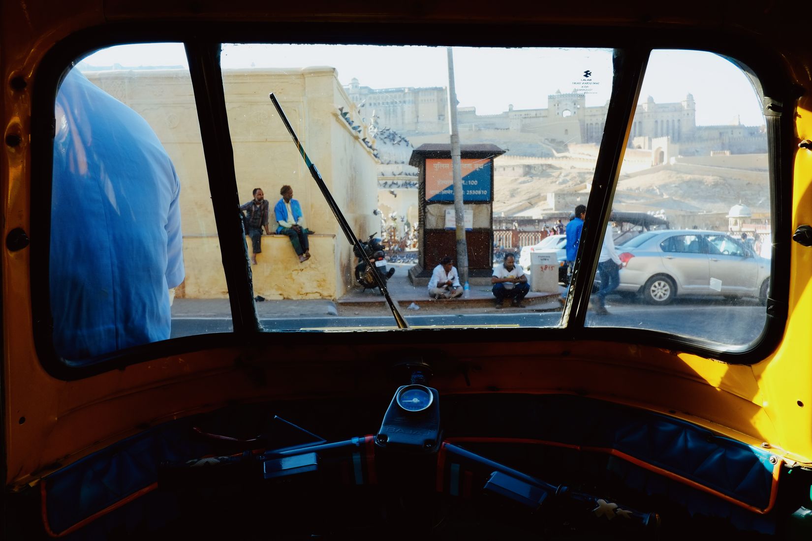 The street framed through the windscreen of an auto-rickshaw, men sitting on a wall opposite, the driver’s blue shirt at the edge