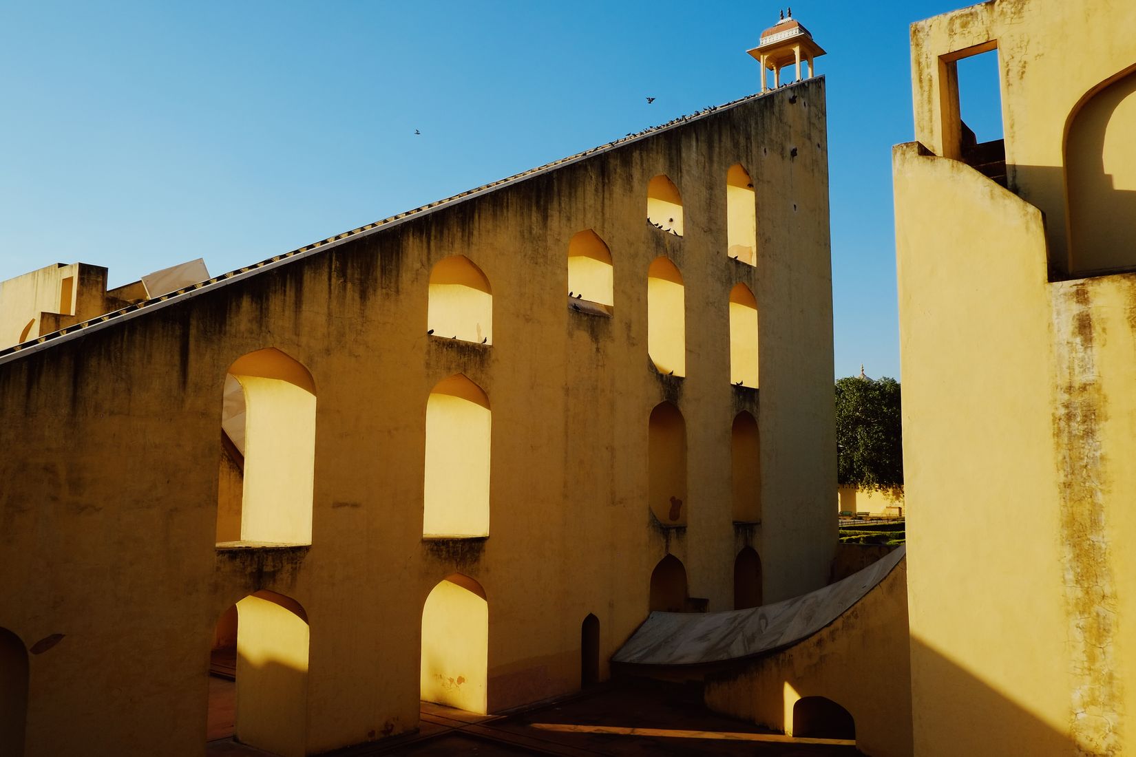 The blank yellow flank of a Jantar Mantar instrument, arched recesses along its length, hard shadow down one side