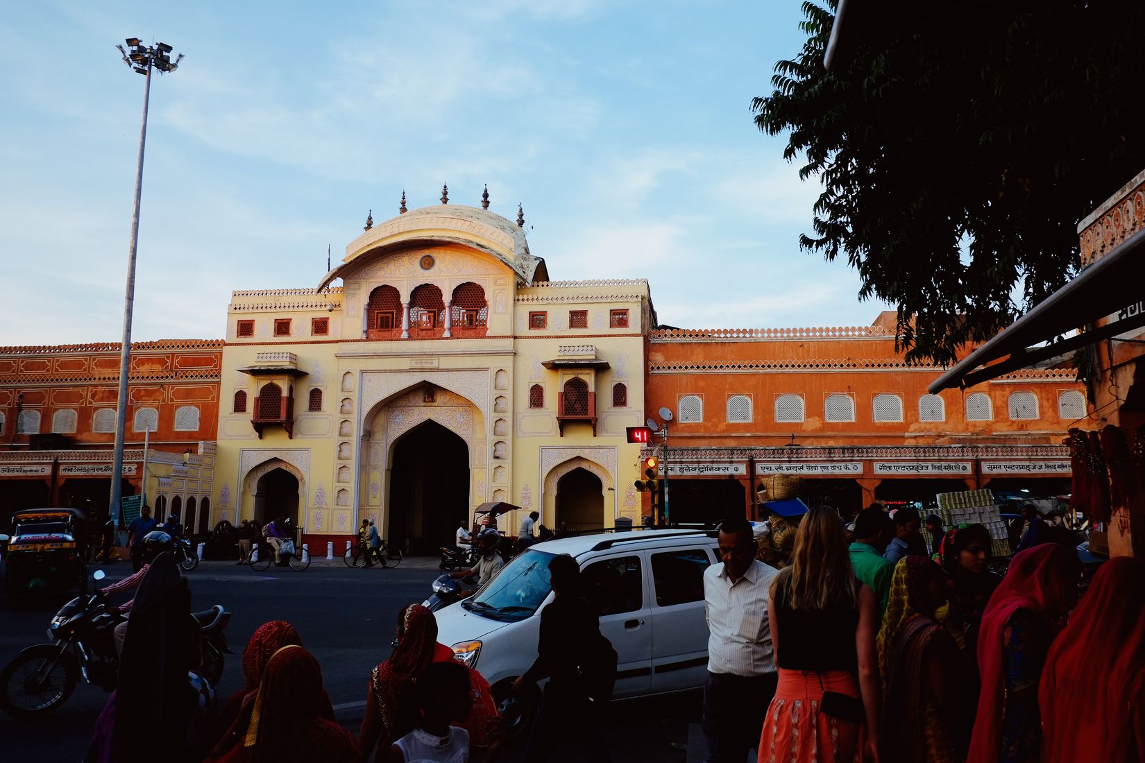 A pink and cream palace gateway across a junction, traffic and a crowd of women in bright saris in front of it