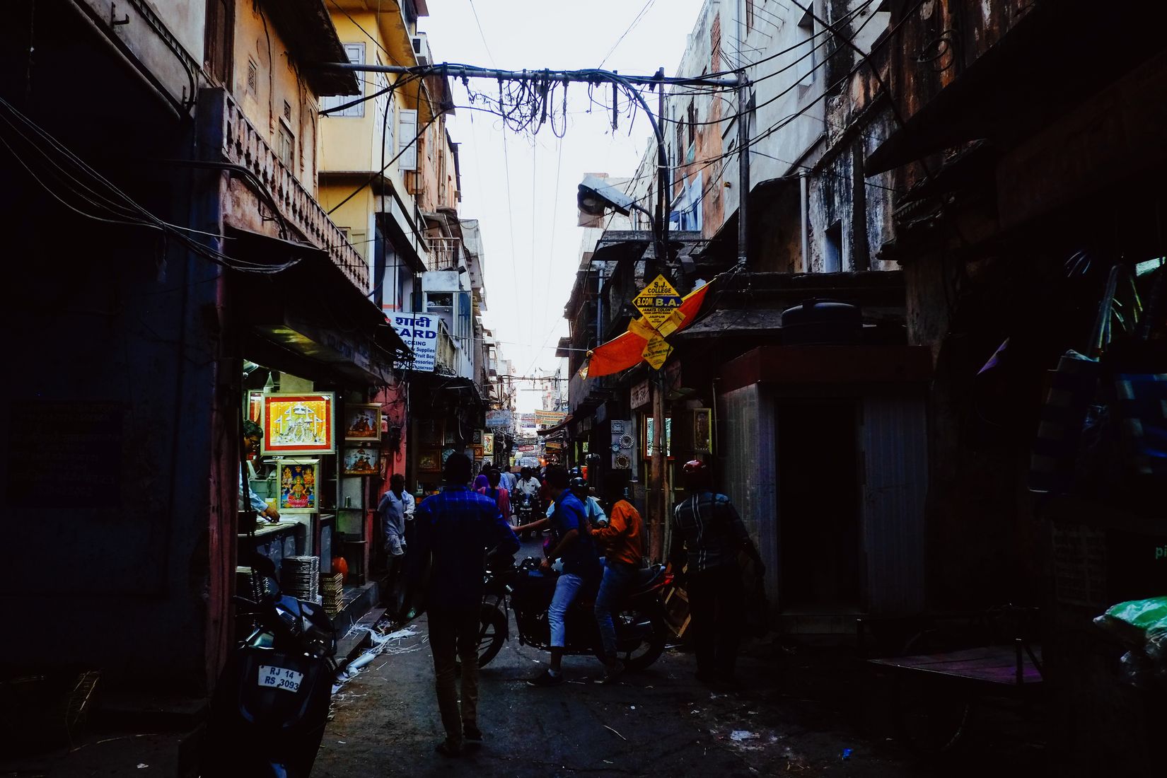 A dim bazaar lane with shops either side, cables slung overhead, figures walking away towards the light at the end