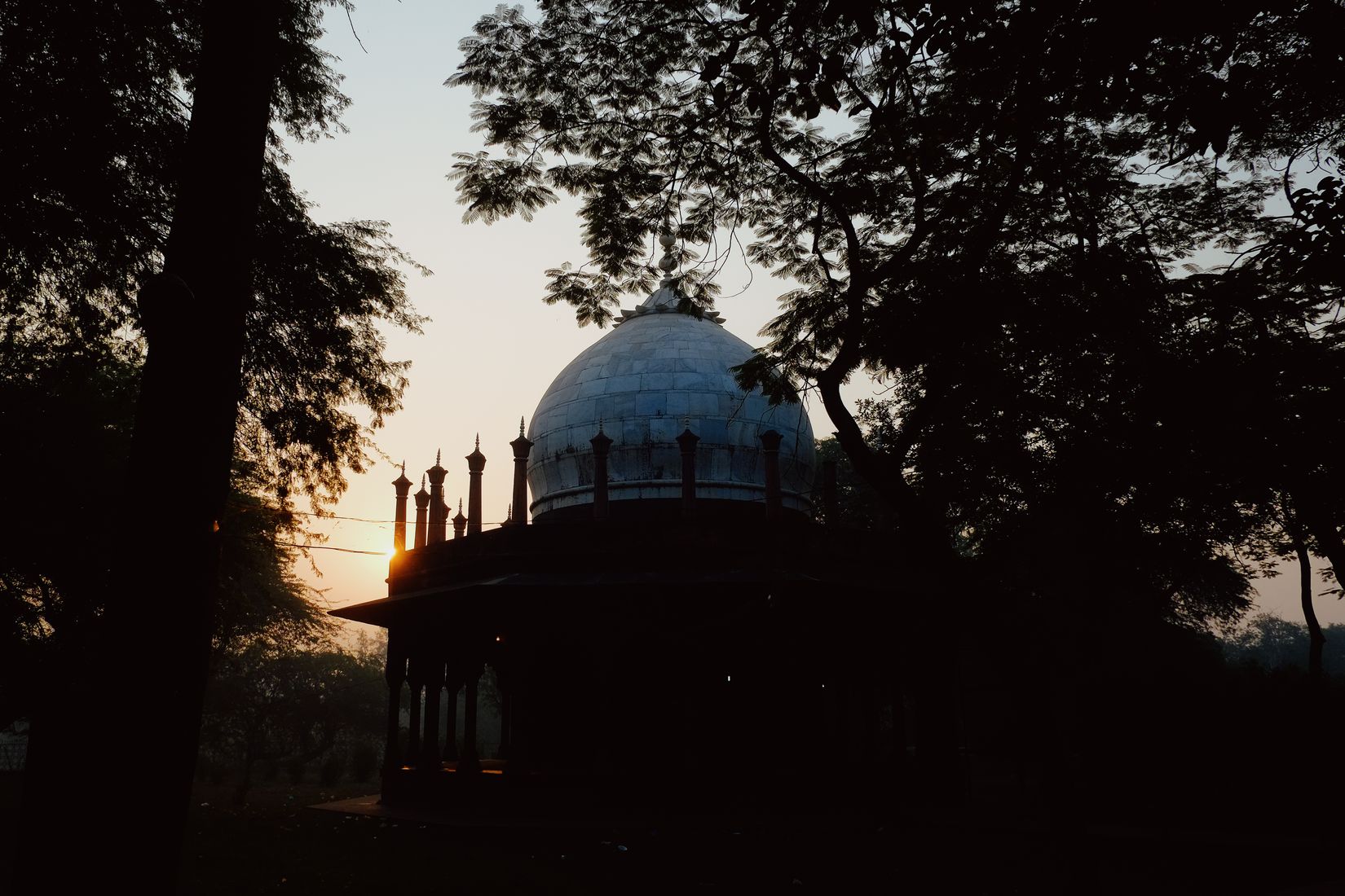 A domed tomb in the trees with the low sun burning through its colonnade