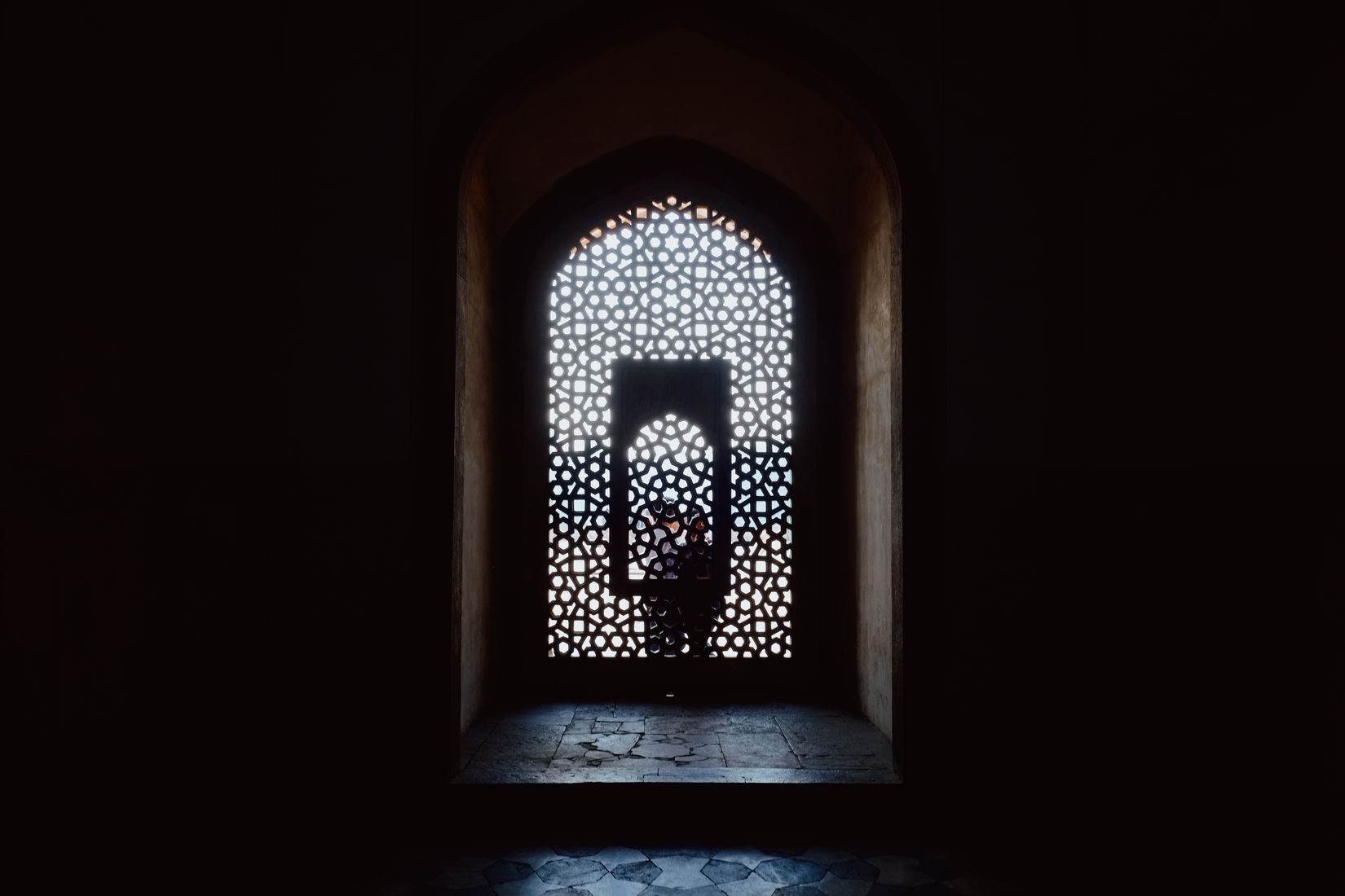 A pierced stone screen in a dark alcove, a figure standing beyond it in the bright arch of the next room