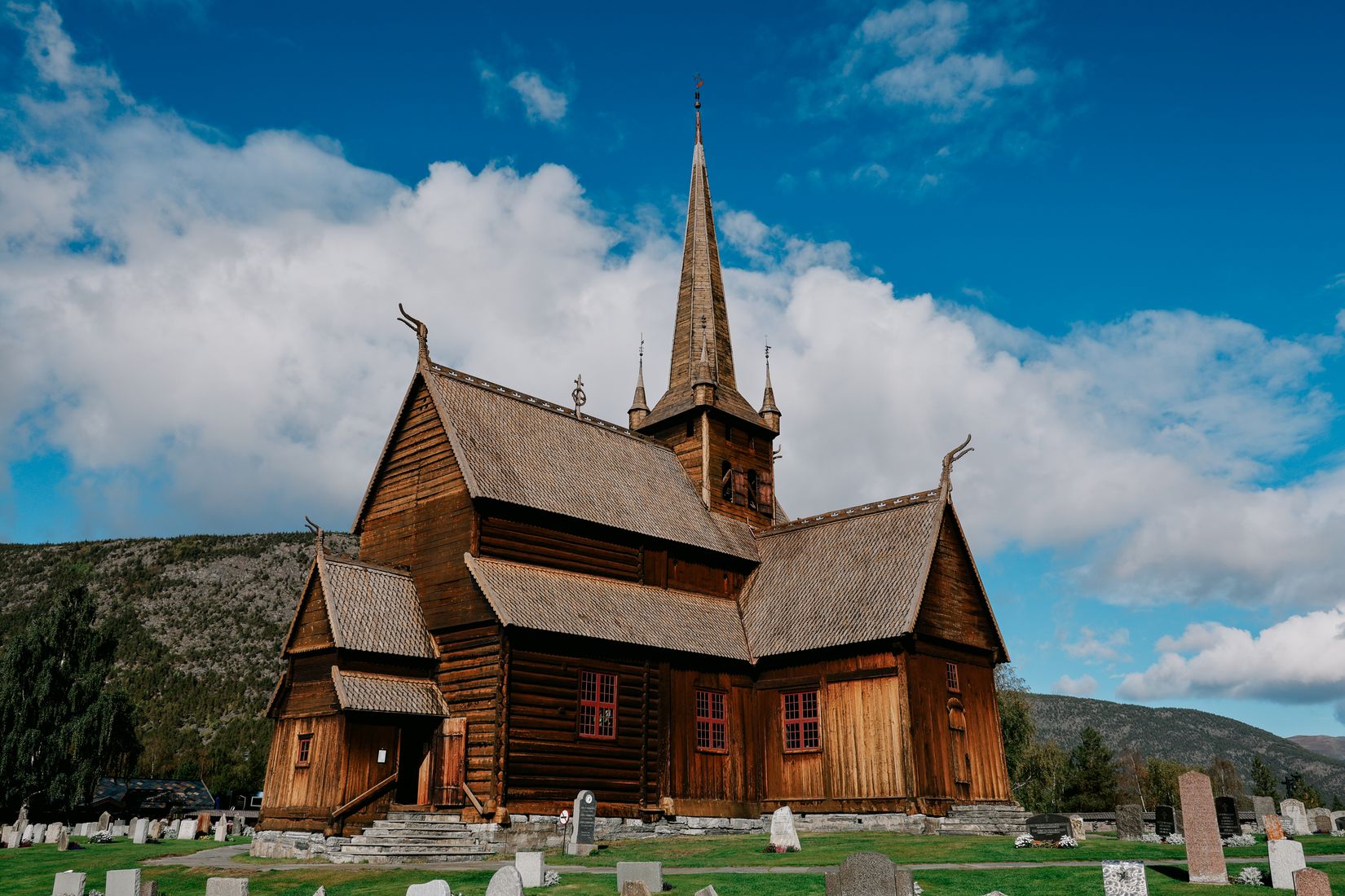 Lom stave church under a blue sky, its tiered wooden roofs and dragon-head gables rising over the gravestones