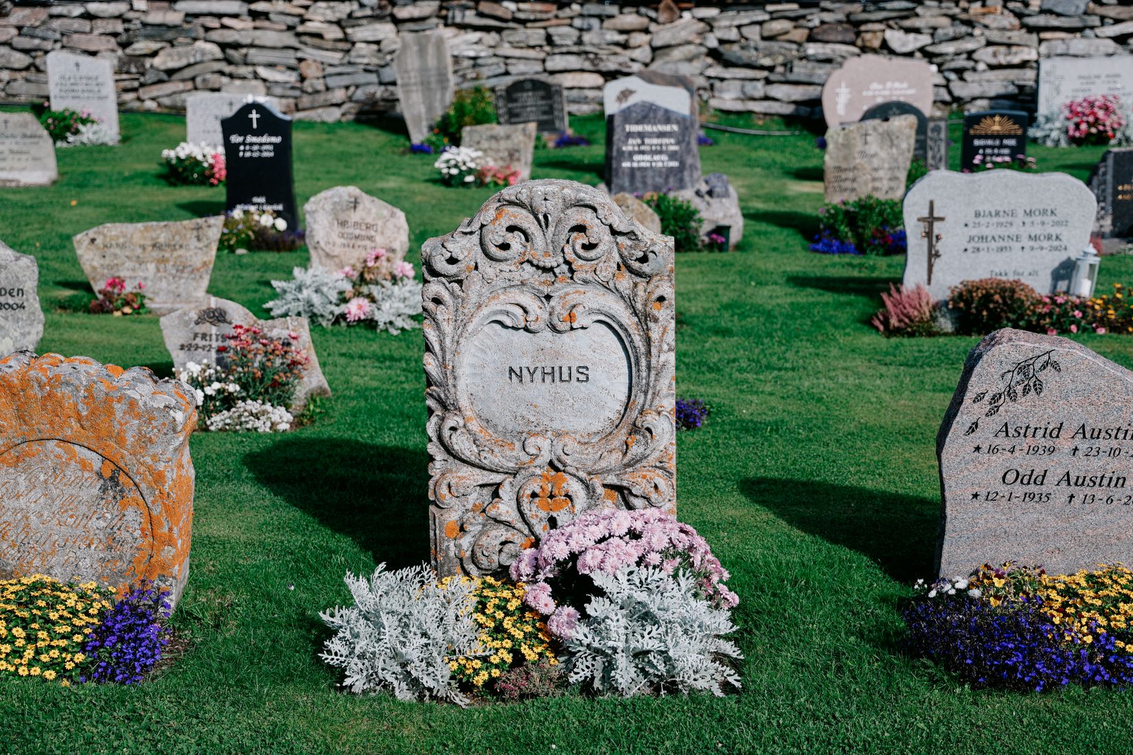 A carved pale headstone reading NYHUS in the churchyard, flowers planted at the foot of every grave