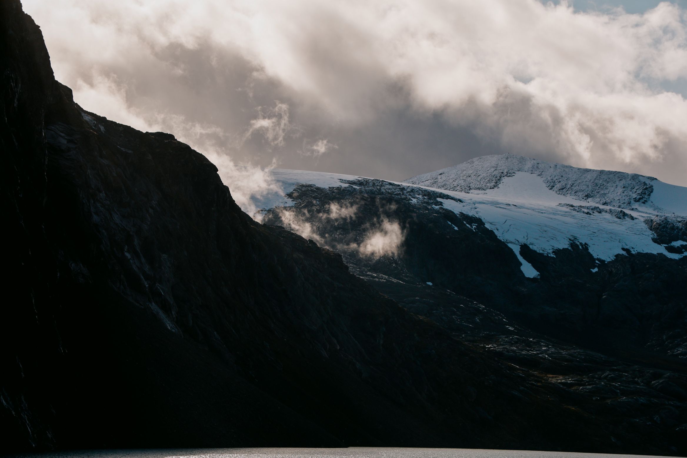 Cloud pouring over a snow-covered plateau above a dark fjord wall, the water pale below