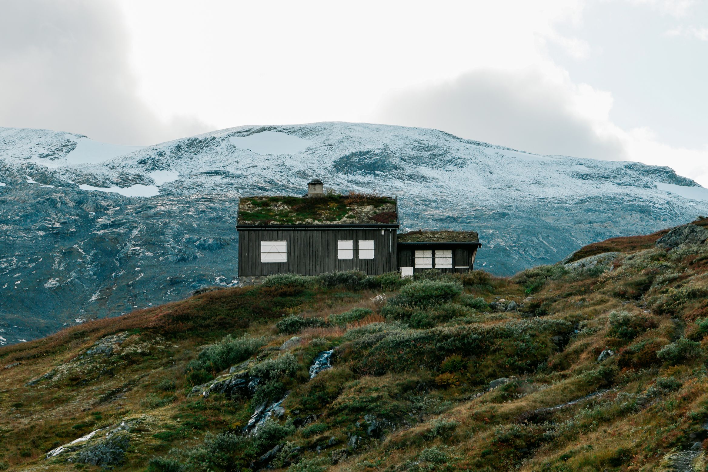 A turf-roofed black timber cabin alone on a ridge, snow-dusted mountains behind it