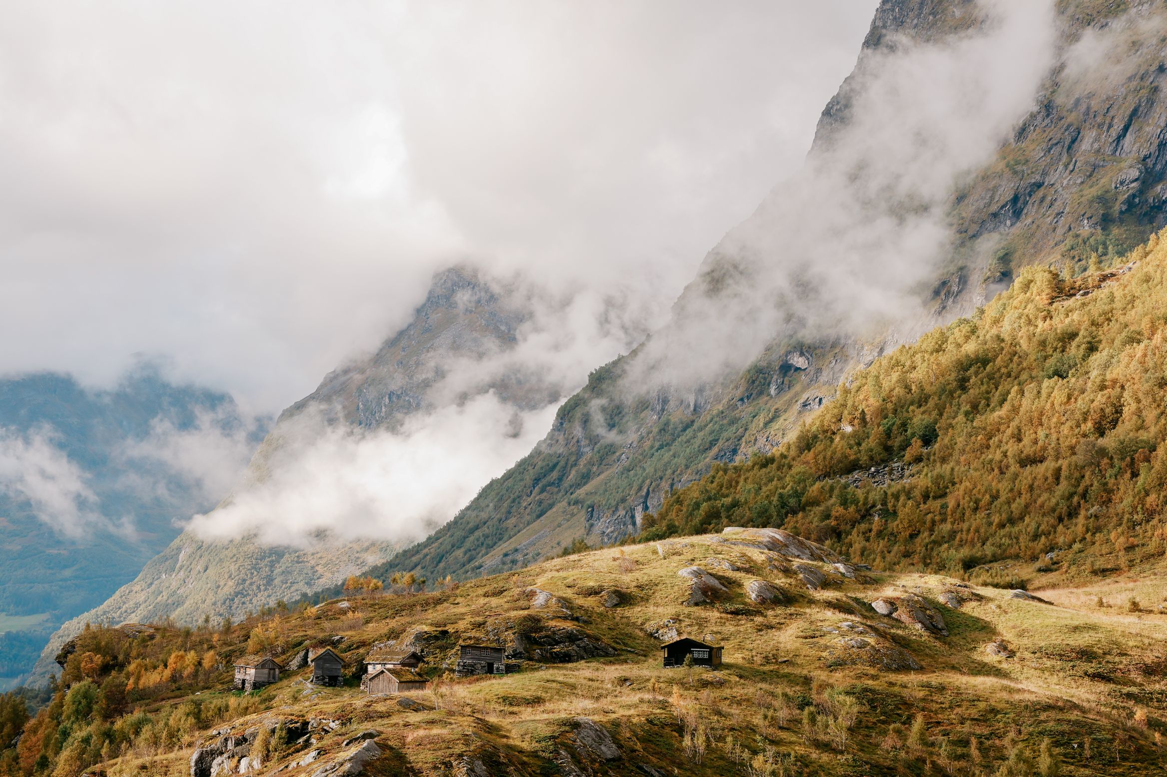 A cluster of turf-roofed farm huts on a high shelf of grass, cloud filling the valley behind