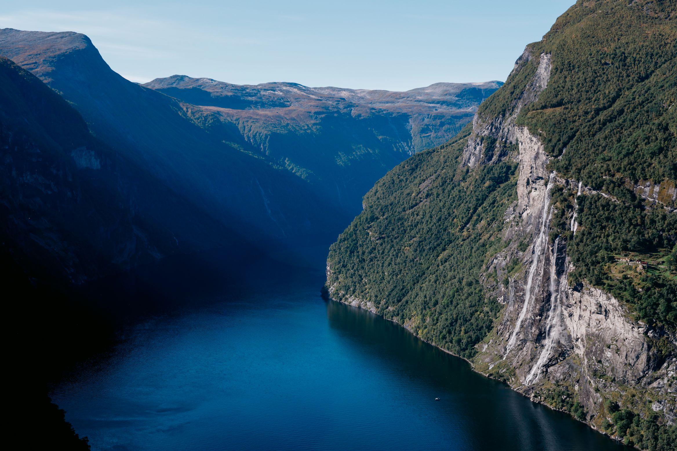 Geirangerfjord from high on the road, deep blue water between sheer walls with waterfalls down the right-hand cliff