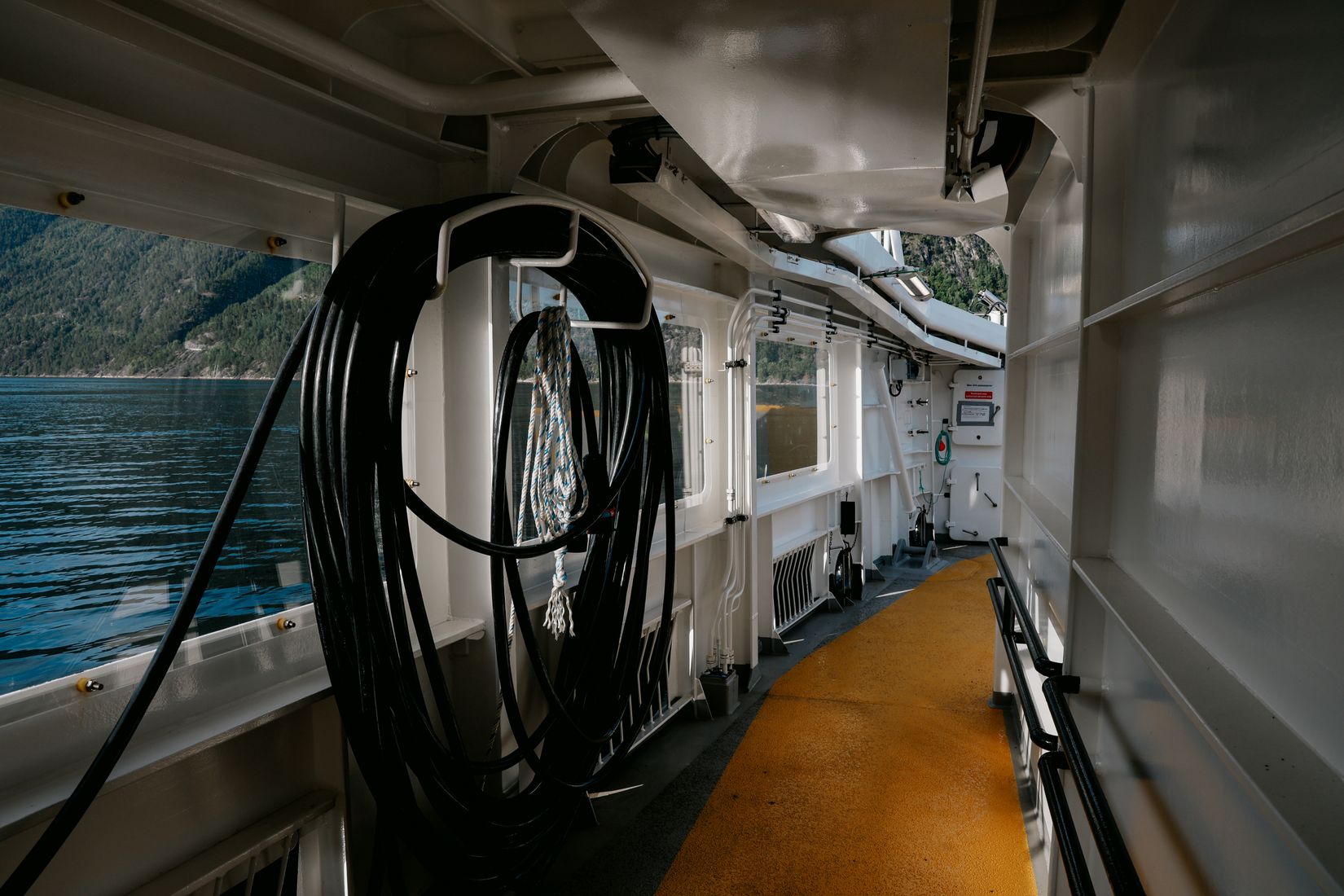 Coiled black hose and a lifebuoy on the ferry side deck, the fjord sliding past the window