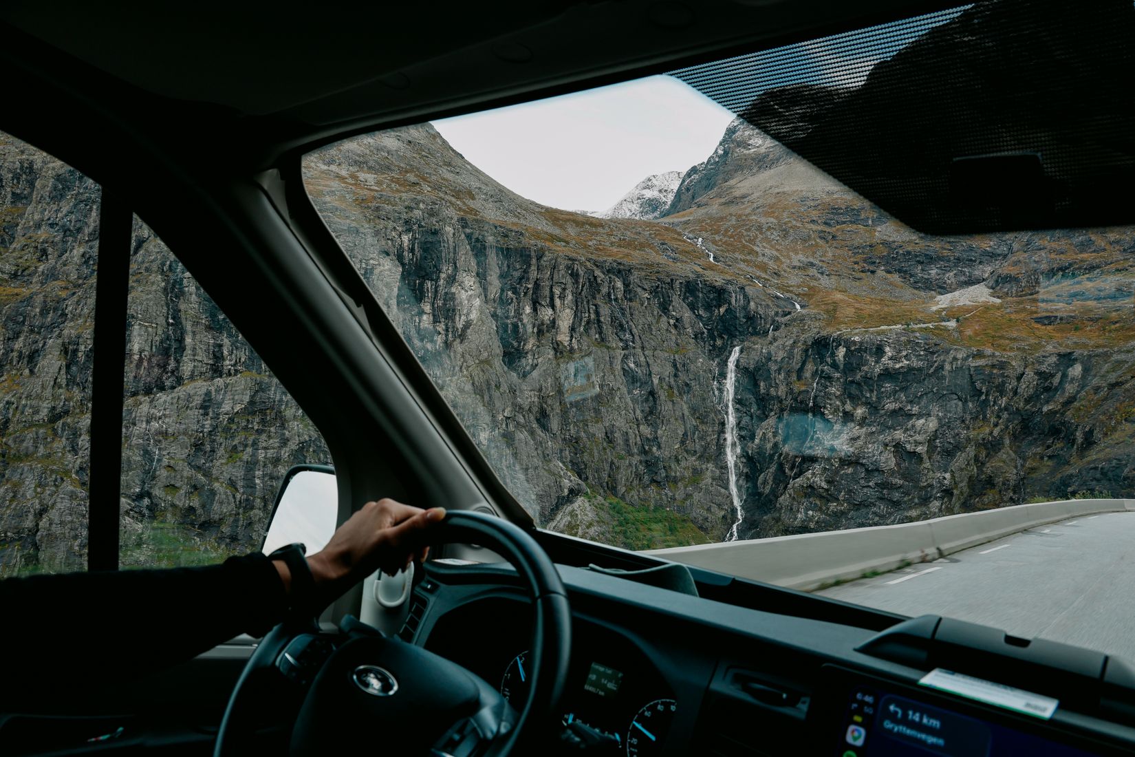 From the passenger seat: a hand on the wheel as the car climbs the Trollstigen hairpins beside a waterfall