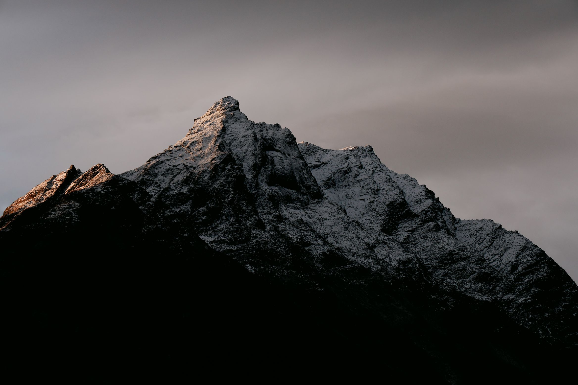 First light catching the snowy tip of a dark peak against a heavy grey sky