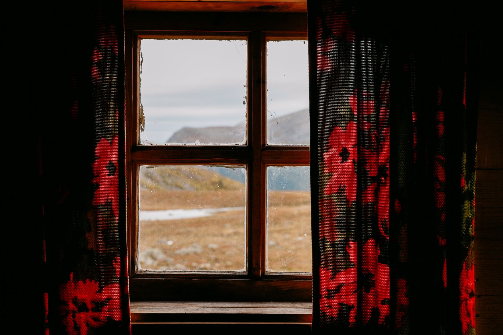 The view out of the hut window through floral red curtains, snow lying on the slope outside