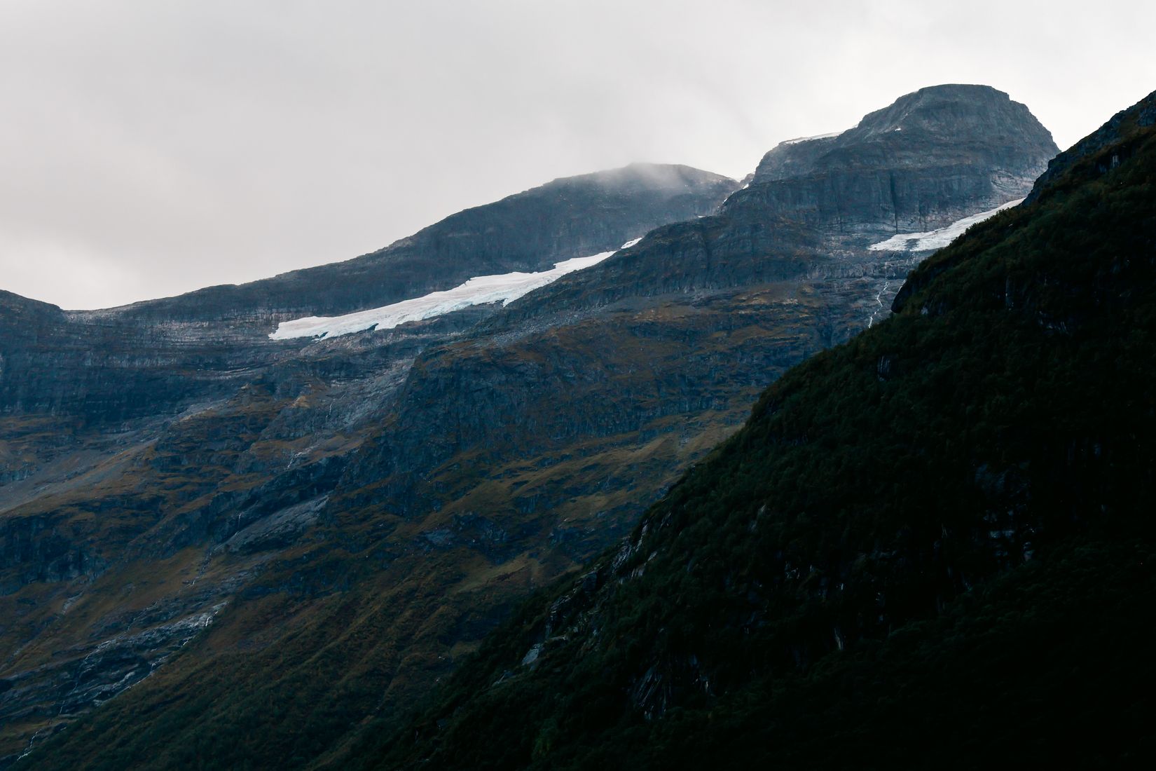 Spindrift blowing off a snow-patched plateau above a steep dark ridge